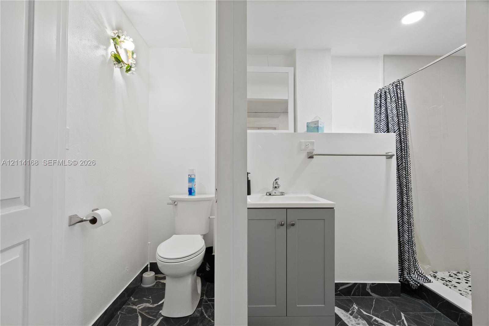 Bathroom featuring a grey vanity with a white countertop, chrome faucet, wall-mounted light fixture, and black veined stone-finish flooring. (Flex space, in-law quarters)