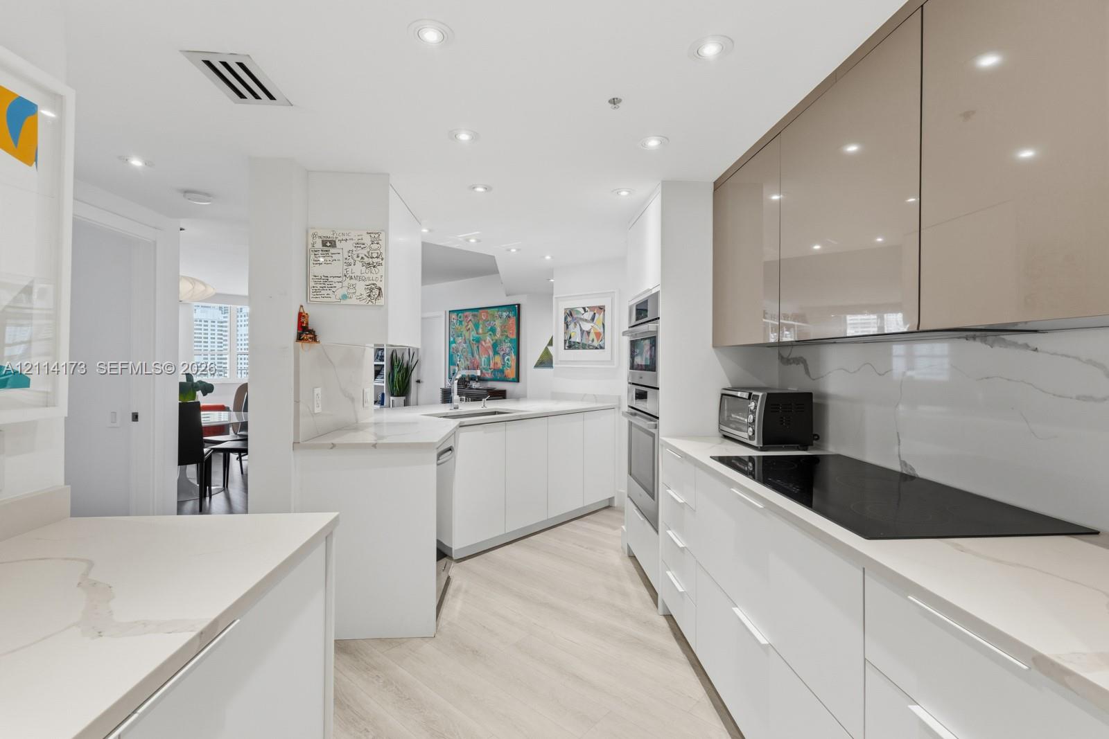 Modern kitchen featuring high-gloss white and metallic cabinetry, stone-finish countertops, full-height stone-finish backsplash, built-in wall oven, and recessed lighting