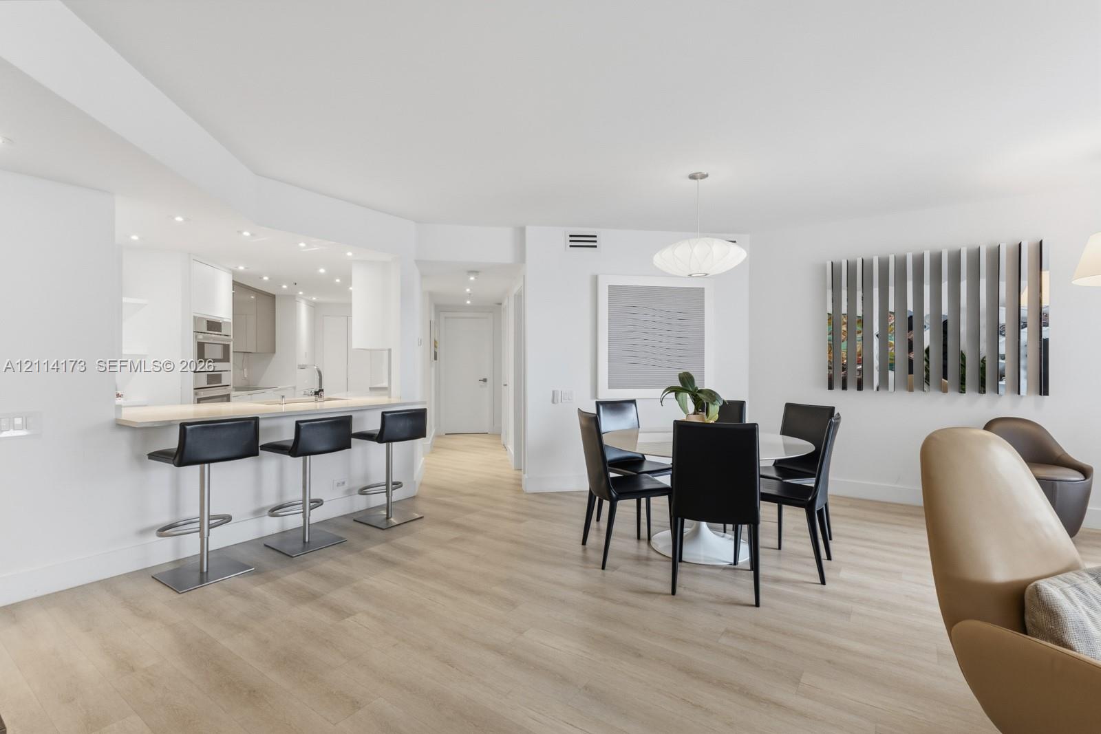 Open-concept space featuring light wood-finish flooring, recessed lighting, built-in kitchen cabinetry with integrated stainless steel appliances, and a quartz-topped breakfast bar