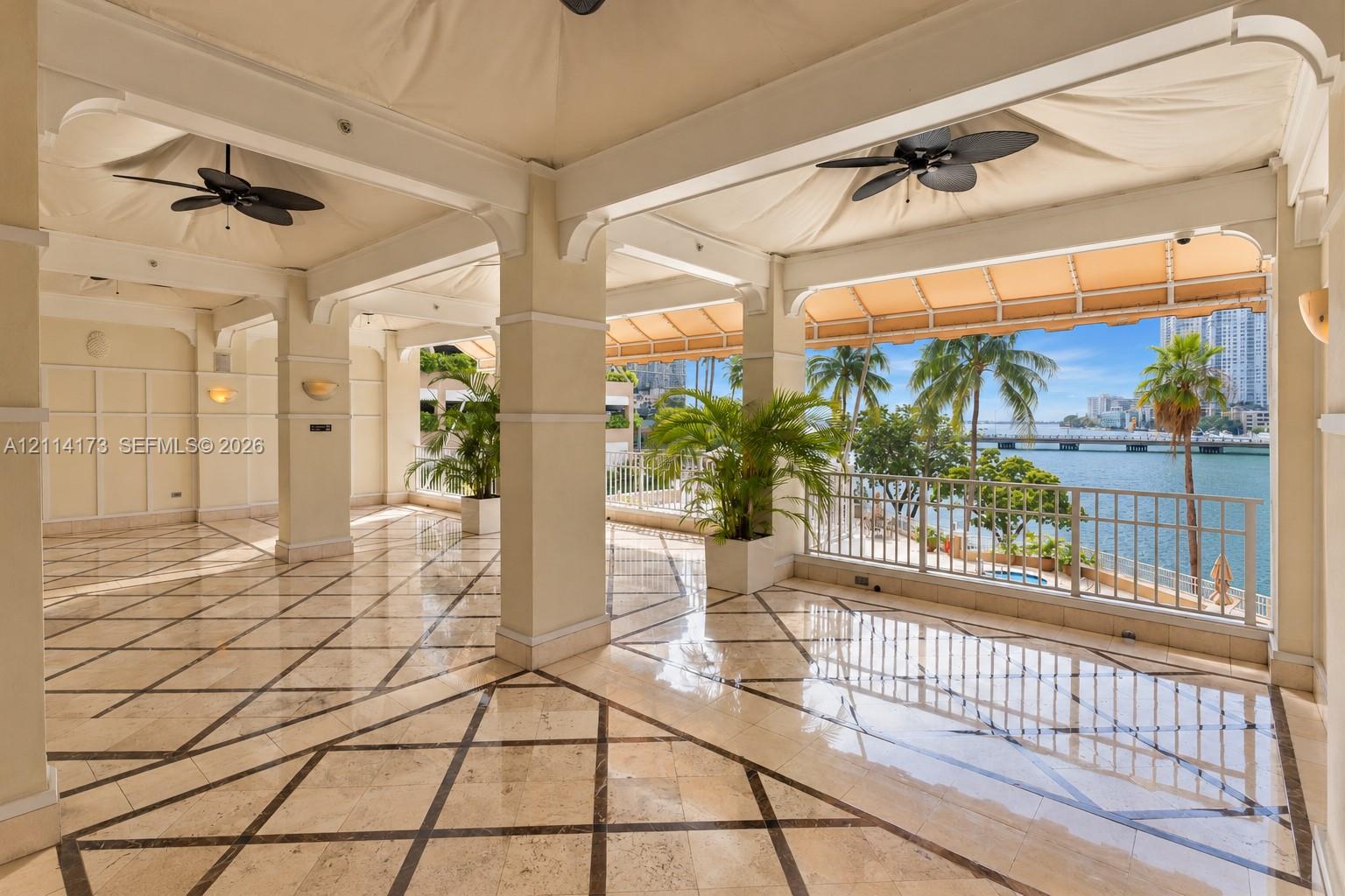 Covered terrace featuring stone tile flooring with decorative inlay, structural columns, ceiling-mounted fans, and metal railings overlooking the water