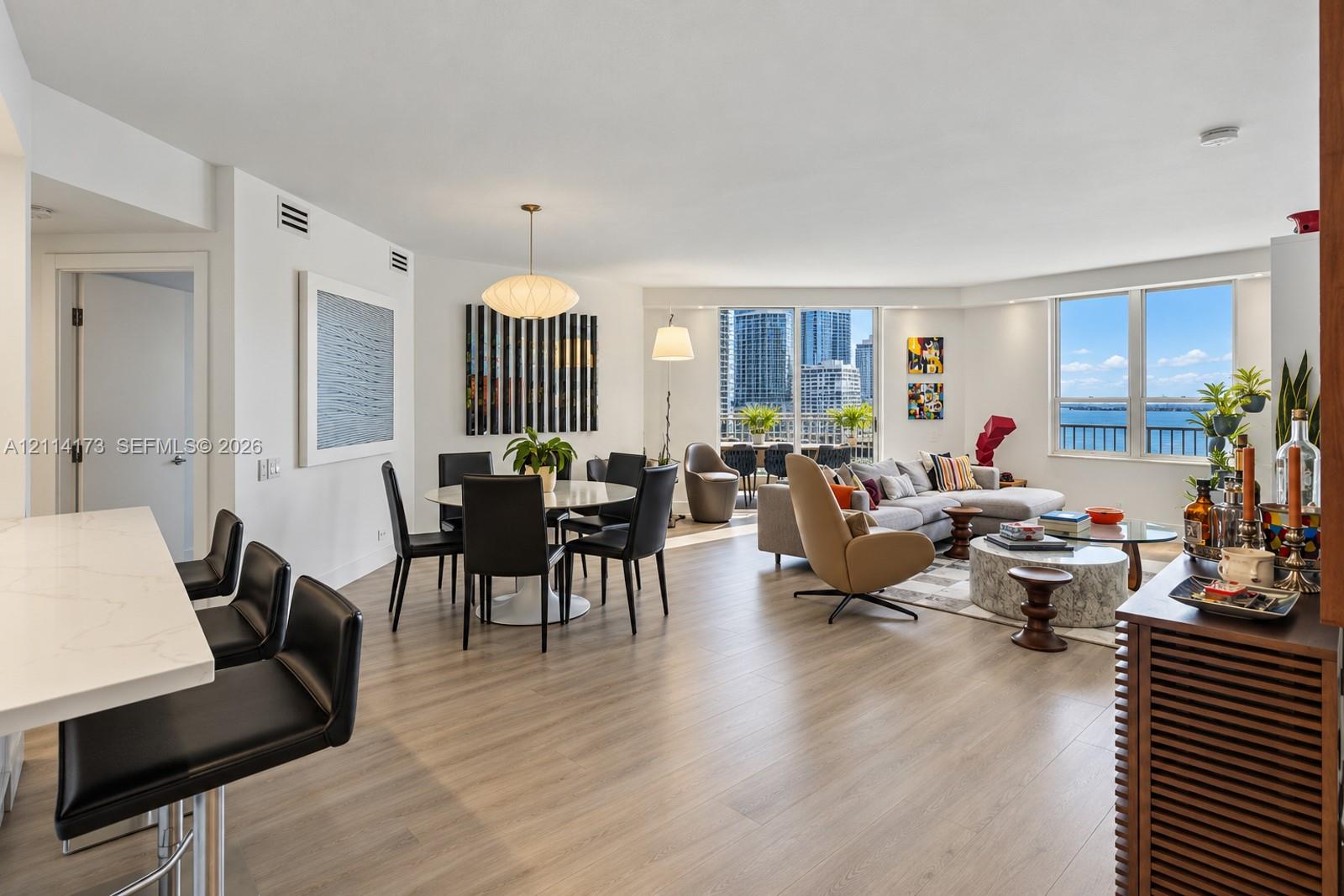 Open-concept main living area featuring light wood-finish flooring, quartz-surfaced kitchen island, recessed lighting, and expansive floor-to-ceiling windows with water and skyline scenery