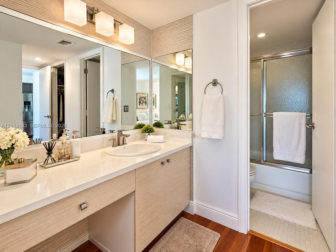 Bathroom featuring a vanity with wood-finish cabinetry, a crisp white countertop, integrated sink, and an expansive wall-to-wall mirror with triple-sconce lighting