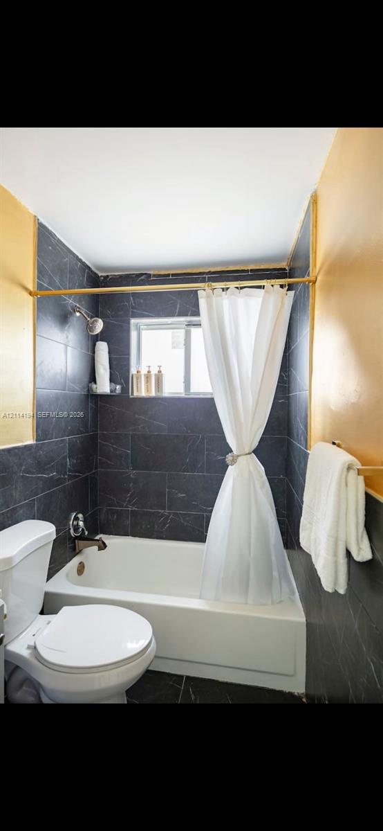 Bathroom featuring dark charcoal stone-finish wall tiling, white porcelain bathtub, metallic finish shower fixtures, and brushed-gold tone hardware