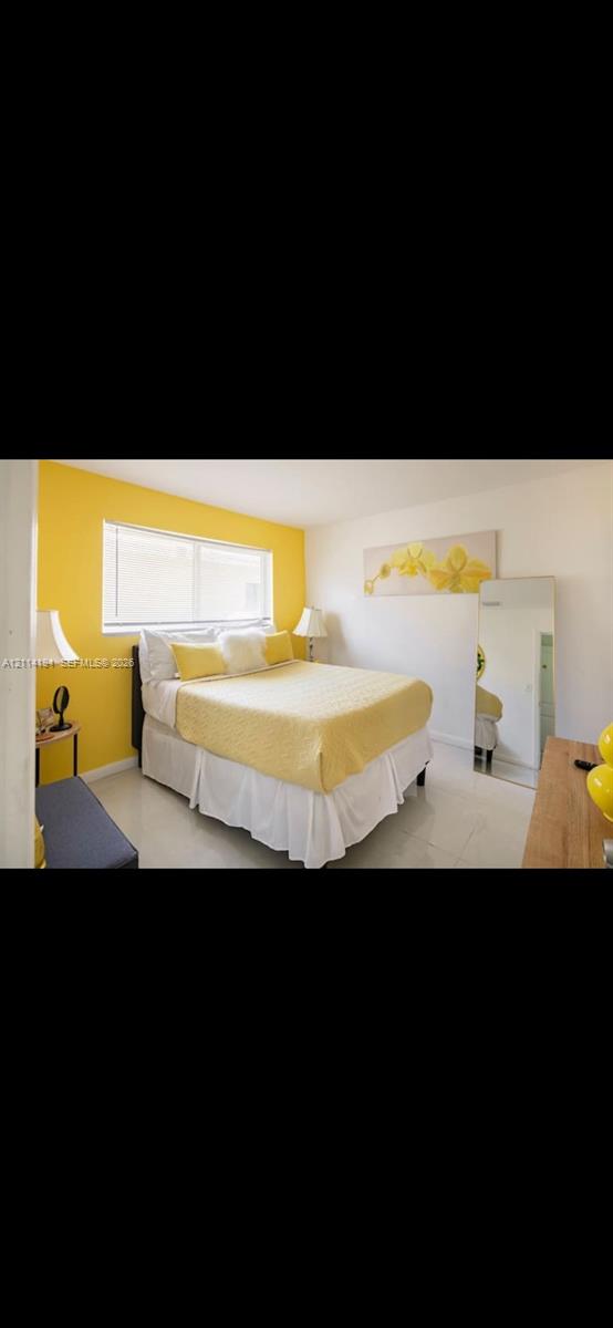 Bedroom featuring light-toned tile flooring, an expansive horizontal window with integrated blinds, and an accent wall