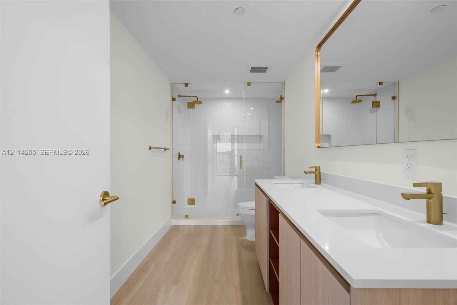 Dual-sink vanity with quartz countertop, light wood-finish cabinetry, brass-toned fixtures, rectangular mirror with metallic frame, and a glass-enclosed shower