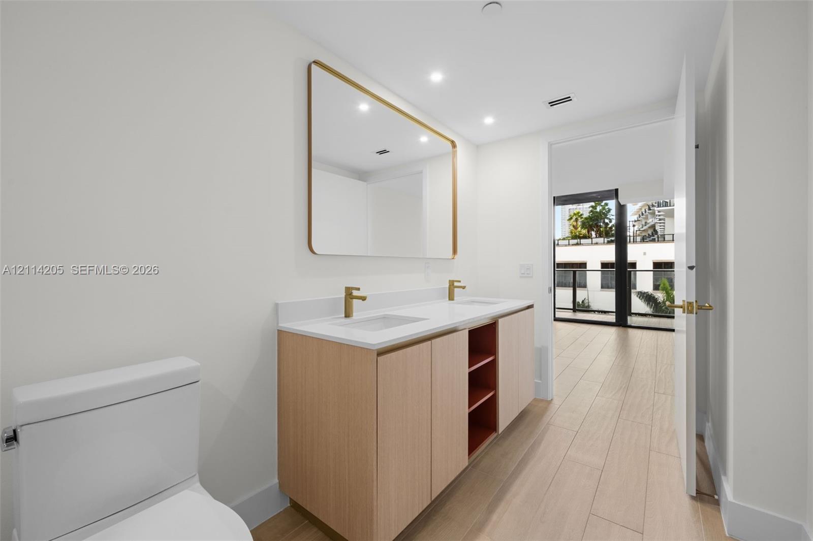 Master bathroom Dual-sink vanity with light wood cabinetry, white stone countertop, rectangular undermount sinks, and gold-tone fixtures, complemented by a large gold-framed wall mirror and wood-finish tile flooring