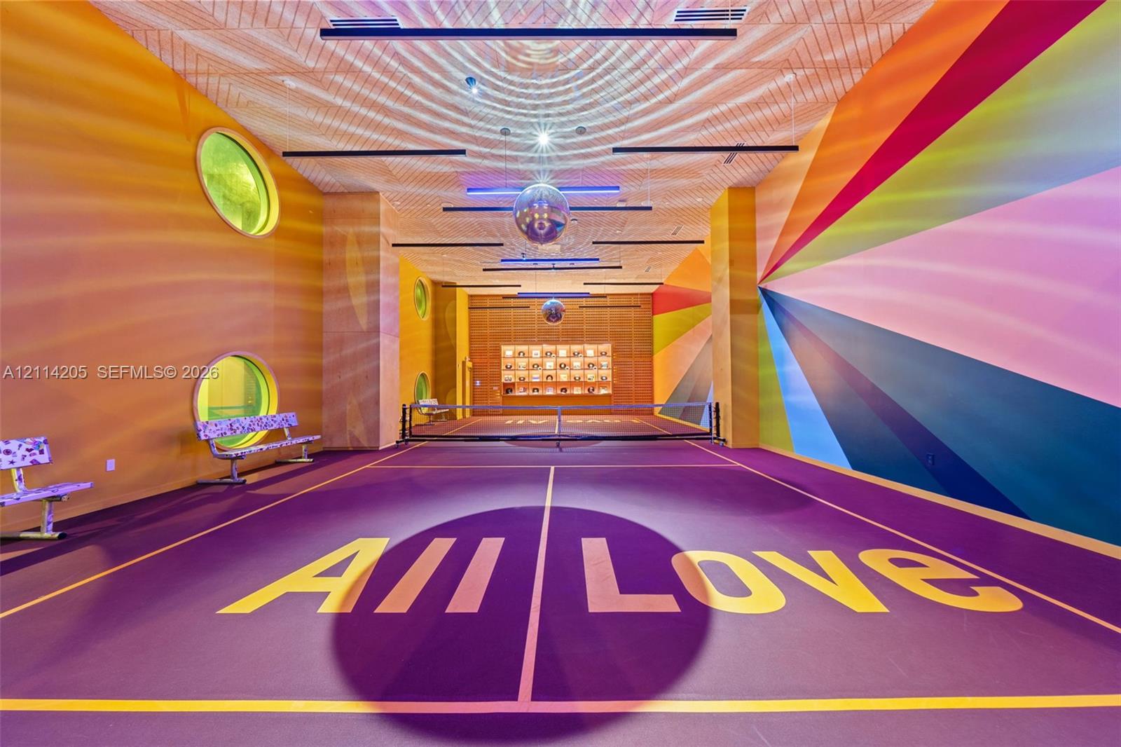 Indoor pickle ball court featuring a high-contrast athletic surface, circular windows, linear ceiling lighting, mirrored spherical pendants, and custom geometric wall murals