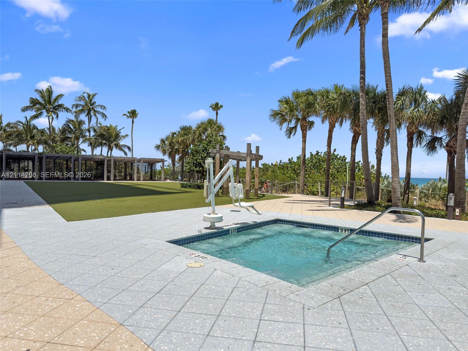 In-ground spa featuring a tiled interior, metal handrails, and a dedicated pool lift, complemented by a large green turf area and paved sun deck