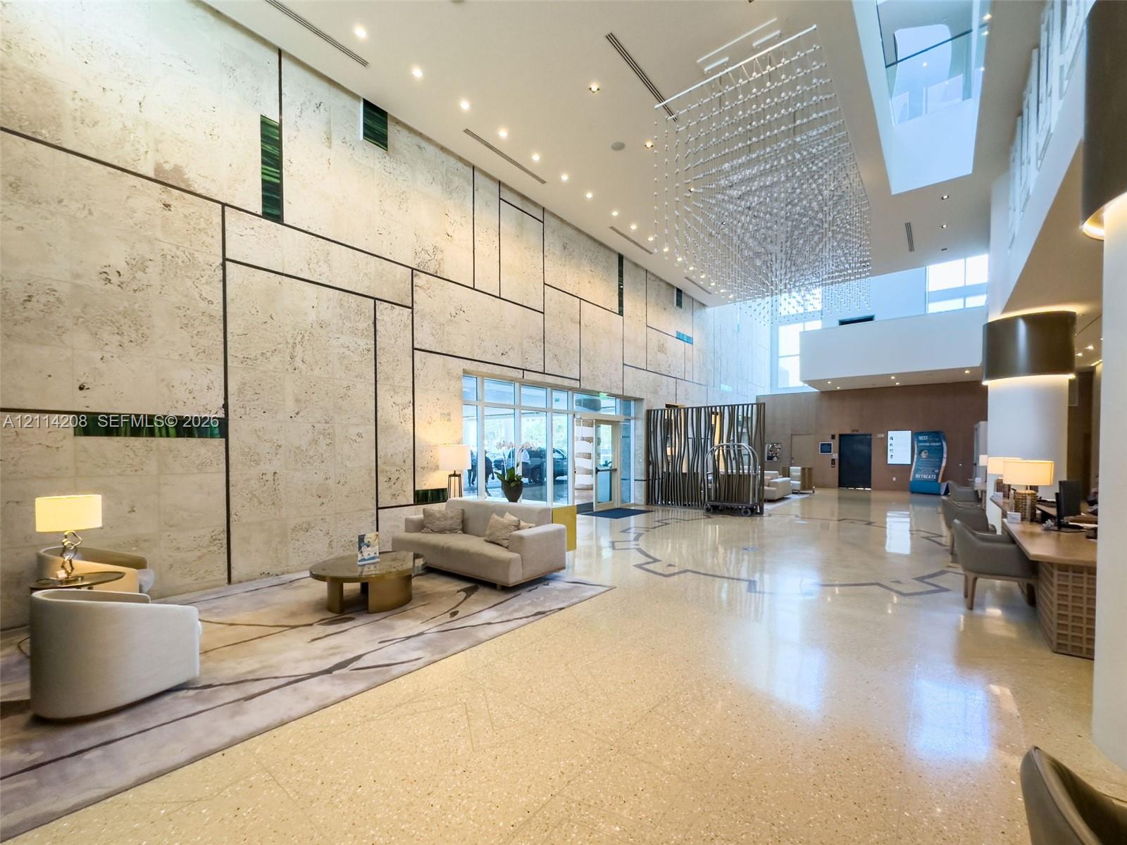 Double-height lobby featuring expansive stone-tiled wall paneling, large-format polished flooring, a tiered crystal chandelier, and recessed lighting