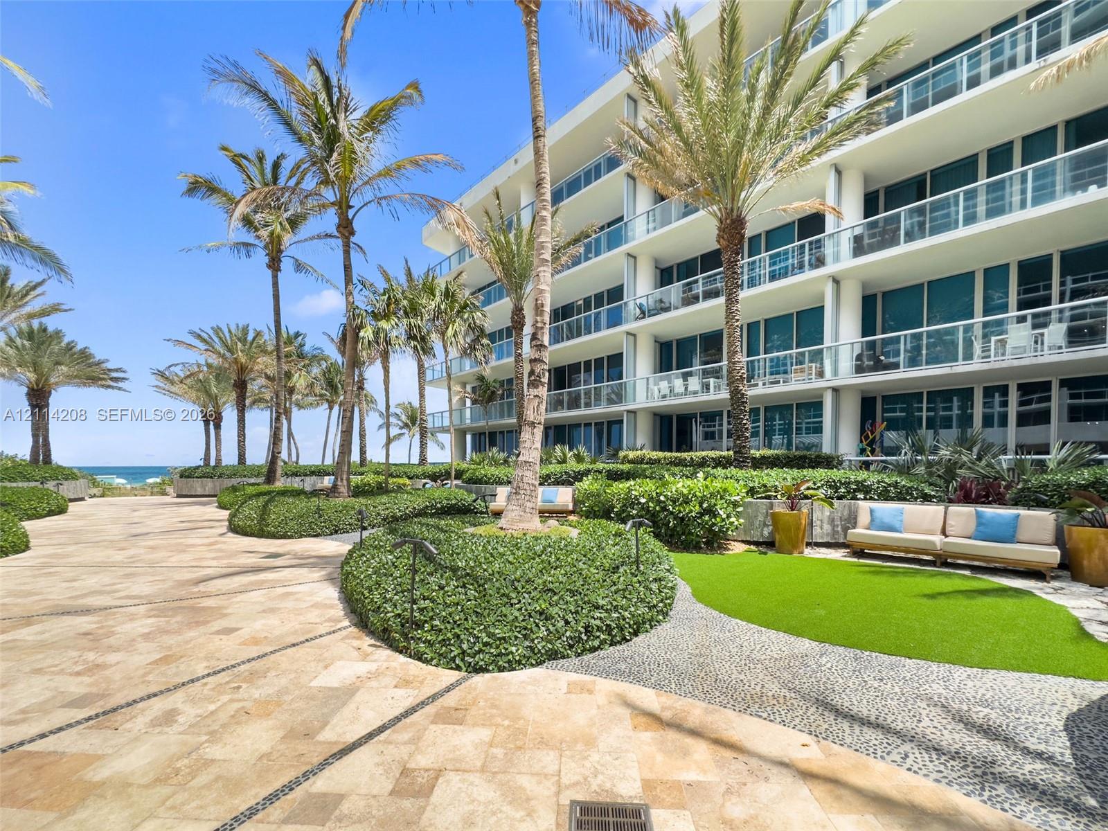 Multi-story building featuring glass-railed balconies, stone-paved walkways, manicured landscaping, and mature palm trees