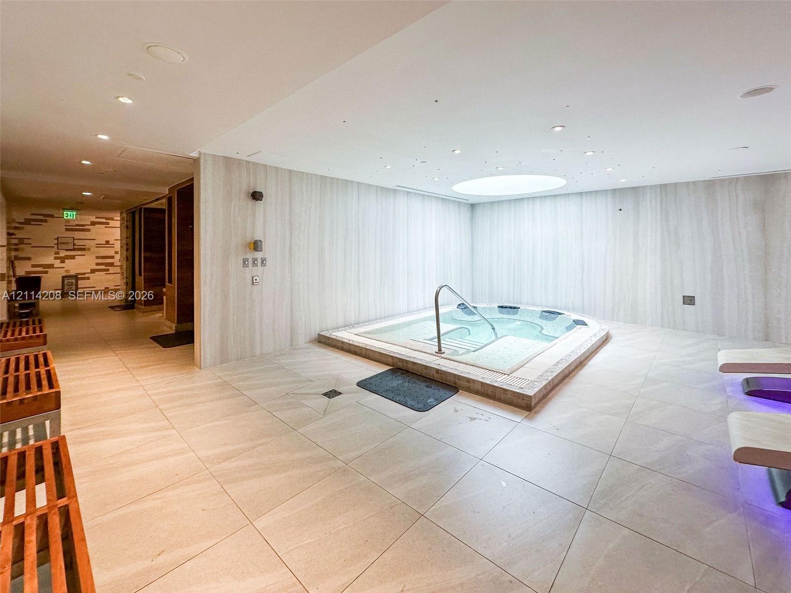 Indoor spa area featuring an integrated jetted hot tub with a metal handrail, large-format tile flooring, light-toned stone-finish wall cladding, and a circular recessed ceiling skylight