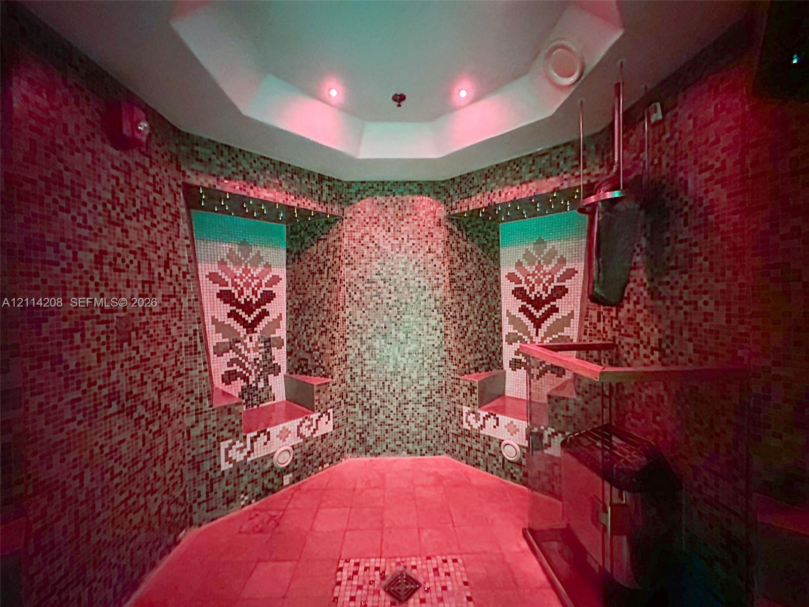 Sauna featuring full-height mosaic tile walls with custom floral inlays, tiered built-in seating, an octagonal ceiling, and a glass-enclosed heater unit