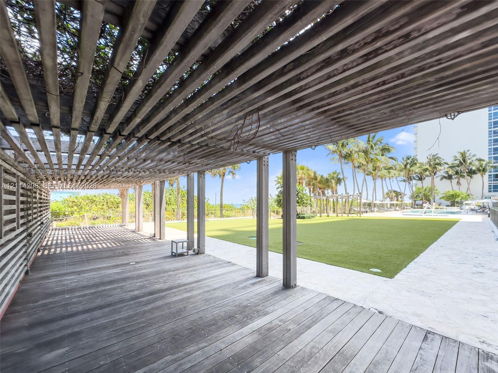Covered wood-plank patio featuring a slatted pergola structure, synthetic turf lawn, stone-paver walkways, and mature palm trees