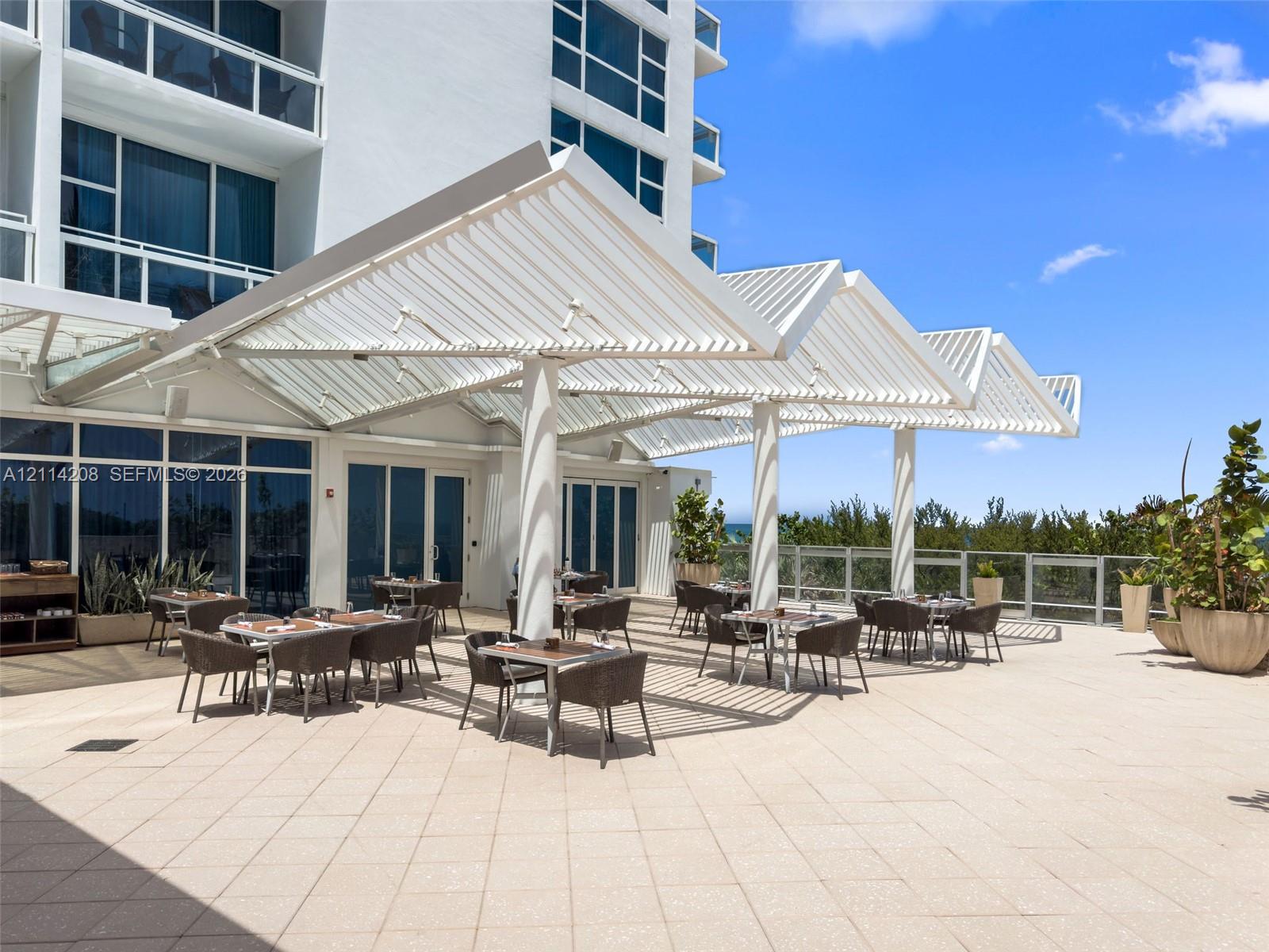 Expansive outdoor terrace featuring light-toned tile flooring, a structured metal louvered canopy, and a glass-paneled perimeter railing