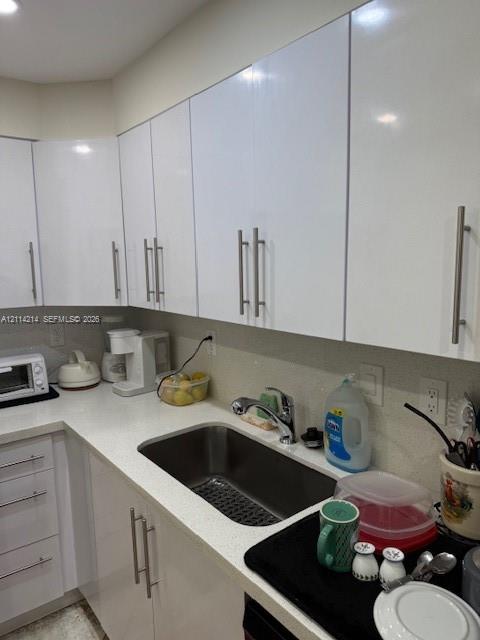 Kitchen featuring white high-gloss cabinetry, light-toned solid surface countertops, a stainless steel undermount sink, and brushed metal hardware