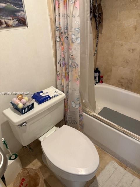 Bathroom featuring a standard white toilet and a built-in bathtub with neutral-toned stone-finish tile surround and a wall-mounted shower fixture