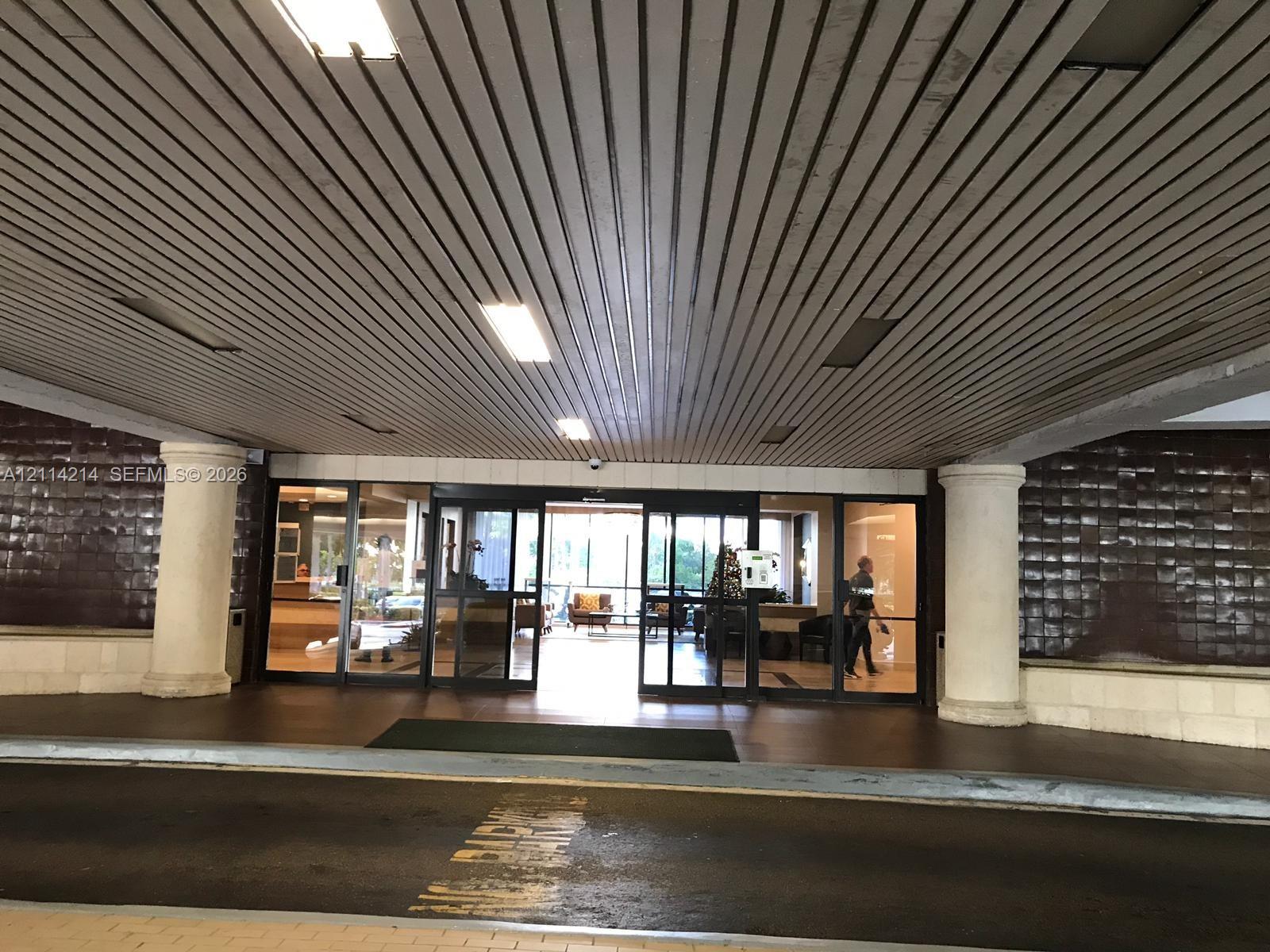 Covered drive-up entrance featuring a slatted ceiling, prominent architectural columns, and expansive glass-paneled sliding entry doors with dark-toned tile exterior siding