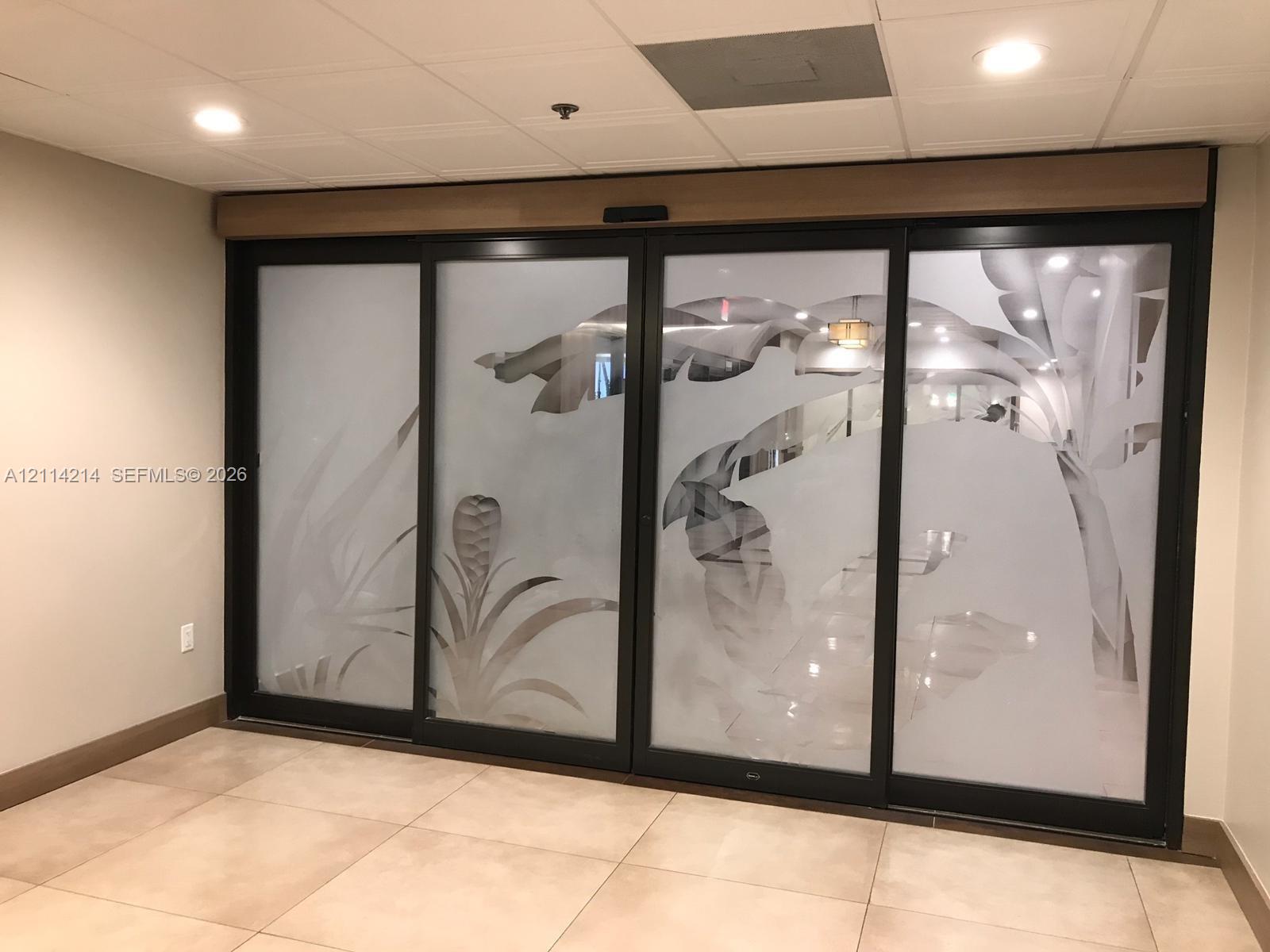 Sliding glass doors featuring black frames and frosted botanical-patterned panels, paired with large-format neutral floor tiling and recessed ceiling lighting