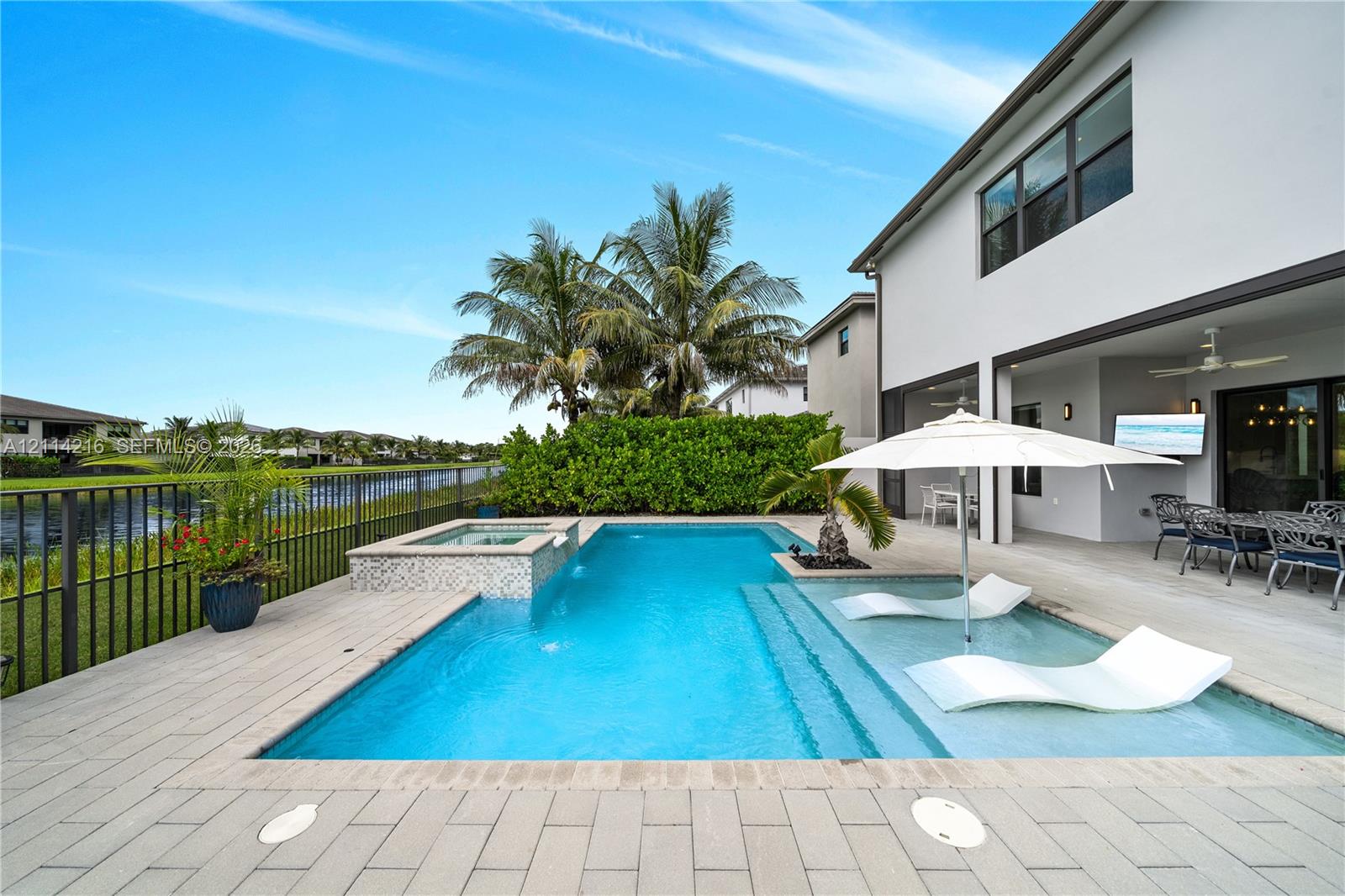 Rear patio featuring a swimming pool with a built-in spa, a tanning ledge, light-toned paver decking, and metal perimeter fencing overlooking a waterway