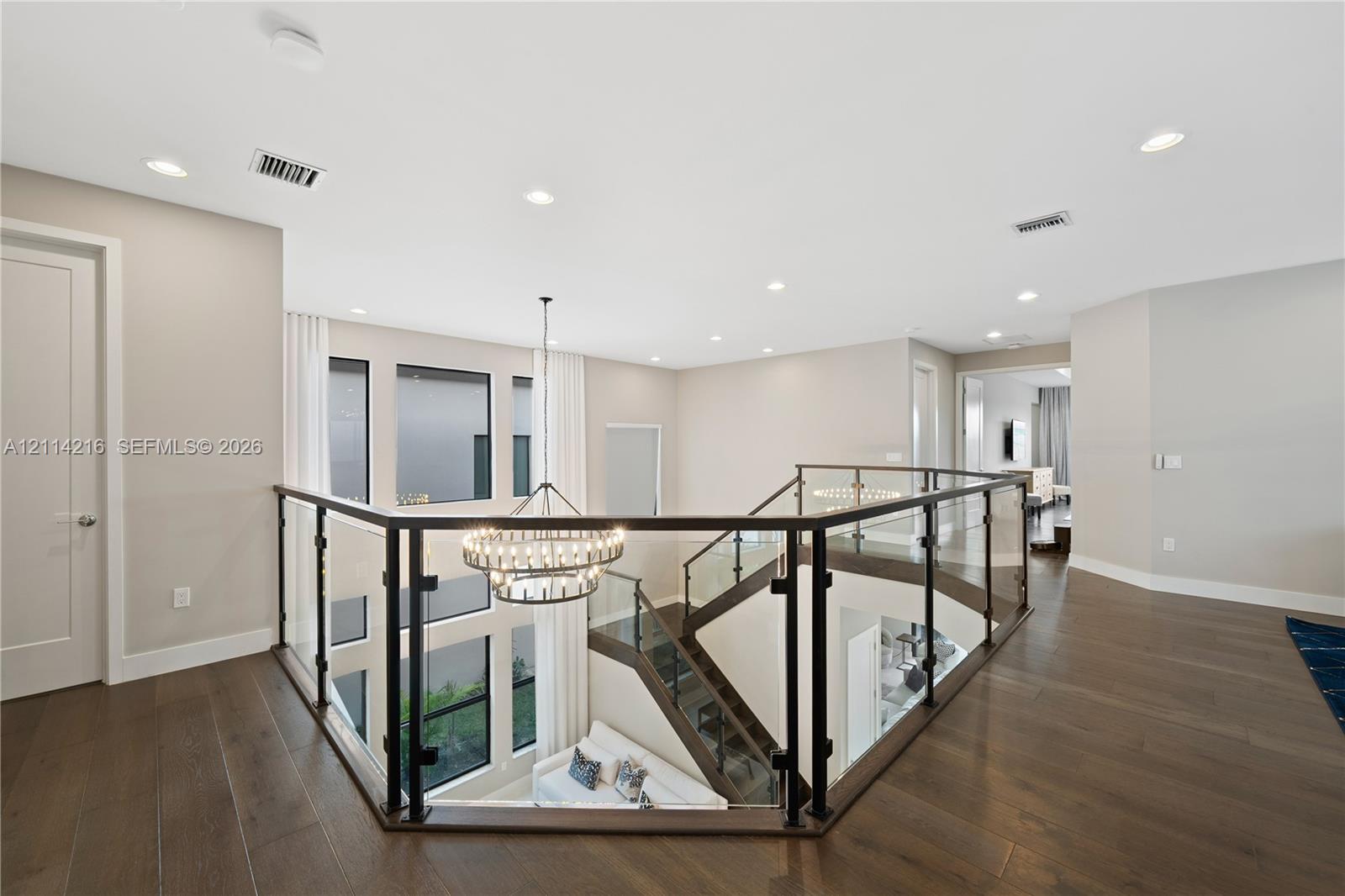 Open mezzanine landing featuring wood-finish flooring, glass panel railing with dark metal framing, recessed lighting, and a multi-tiered circular chandelier
