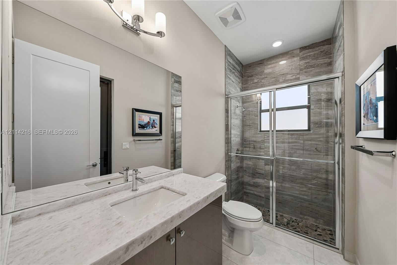 Vanity with light-toned stone-finish countertop and integrated rectangular basin, full-width mirror, glass-enclosed shower with horizontal textured tile surround, and chrome hardware