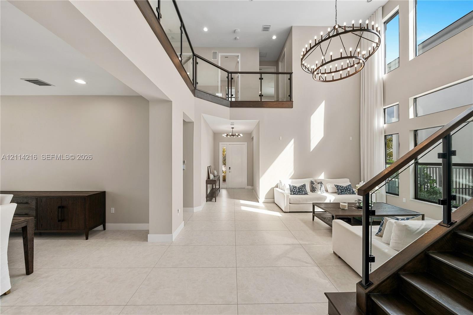 Double-height ceiling, open-concept floor plan with large-format tile flooring, staircase with dark wood treads and glass railings, multi-tiered circular chandelier, and expansive vertical windows