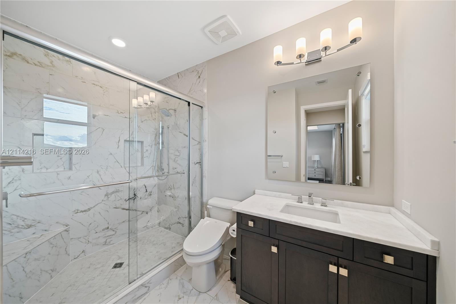 Bathroom featuring a glass-enclosed walk-in shower with stone-patterned tile, dark wood-finish cabinetry, a white countertop with an integrated rectangular sink, and a wall-mounted vanity mirror with a four-light fixture