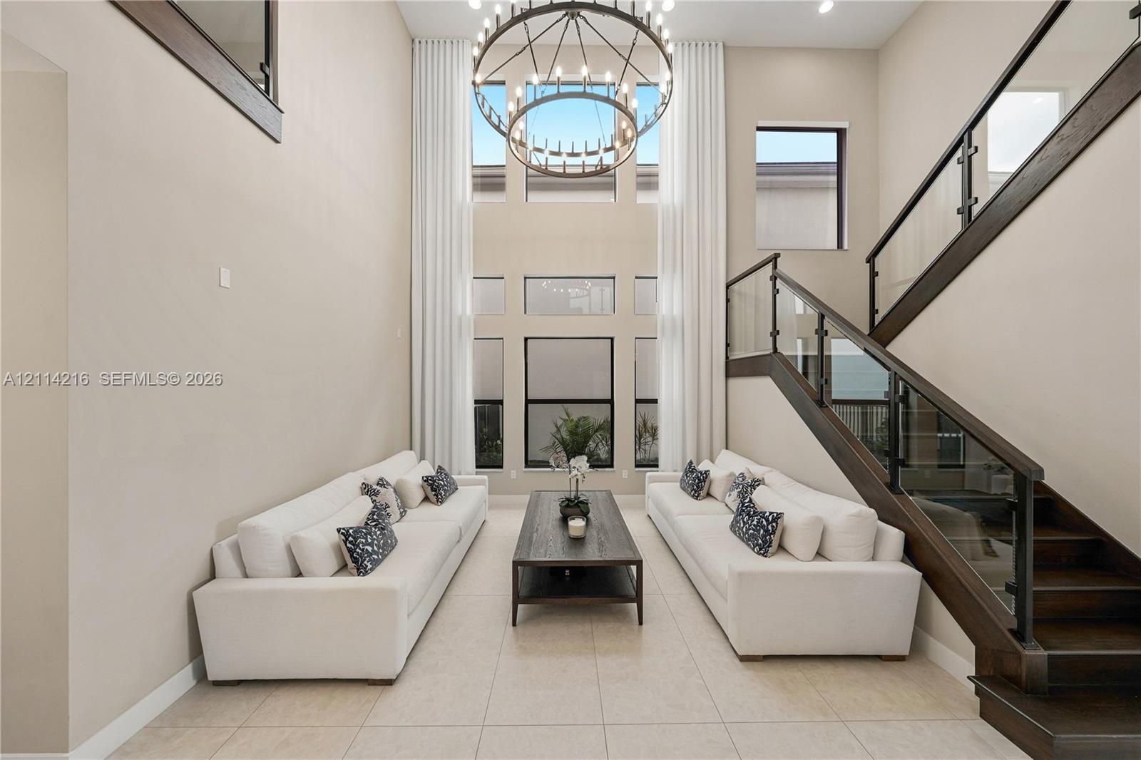 Two-story height ceiling with expansive windows, custom iron-frame chandelier, and staircase featuring dark wood finishes and glass railing panels