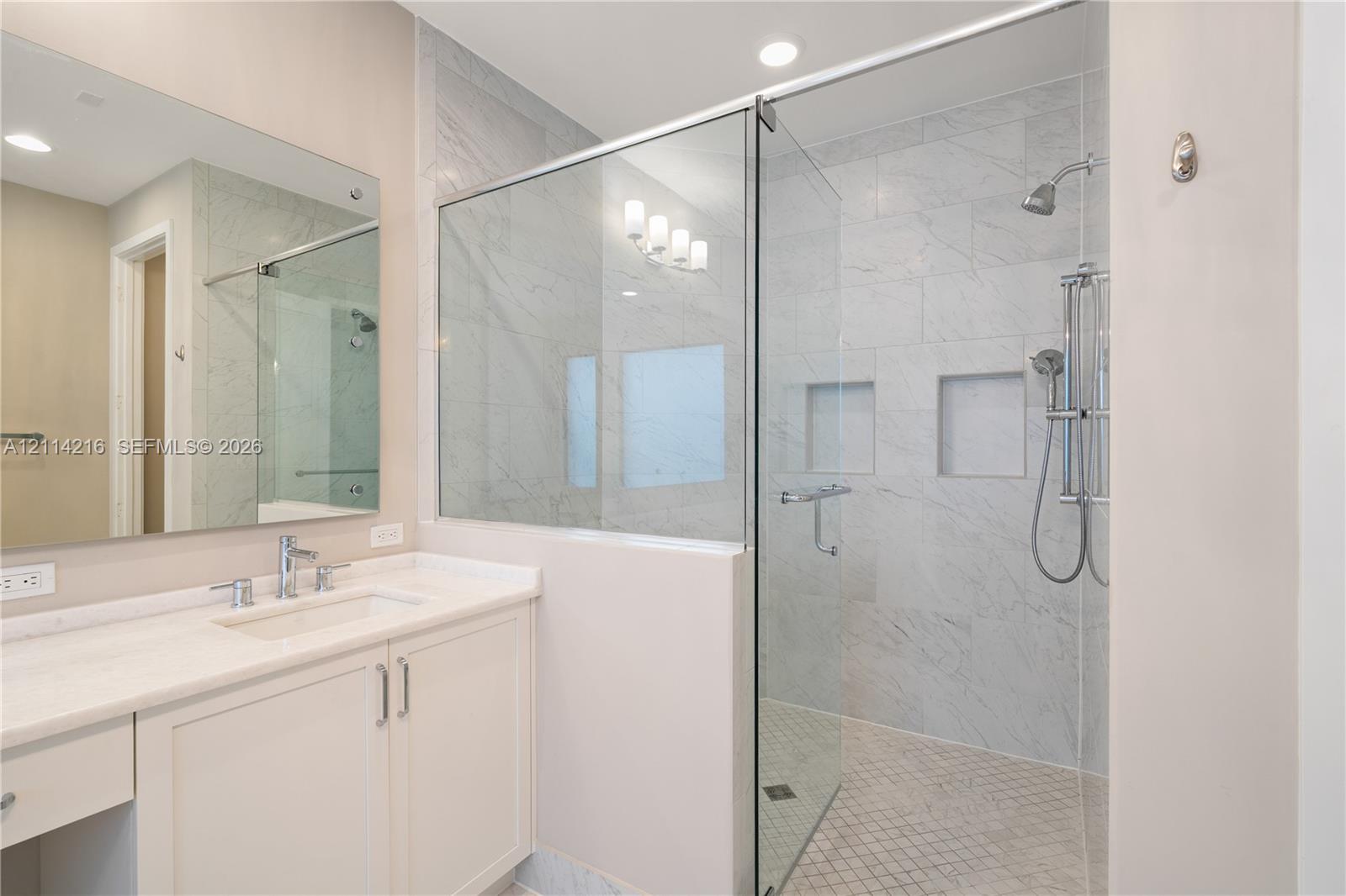 Bathroom featuring a vanity with a light-toned countertop, undermount sink, and integrated cabinetry, paired with a walk-in shower enclosed by glass panels, stone-finish wall tiling, dual recessed niches, and a handheld shower fixture
