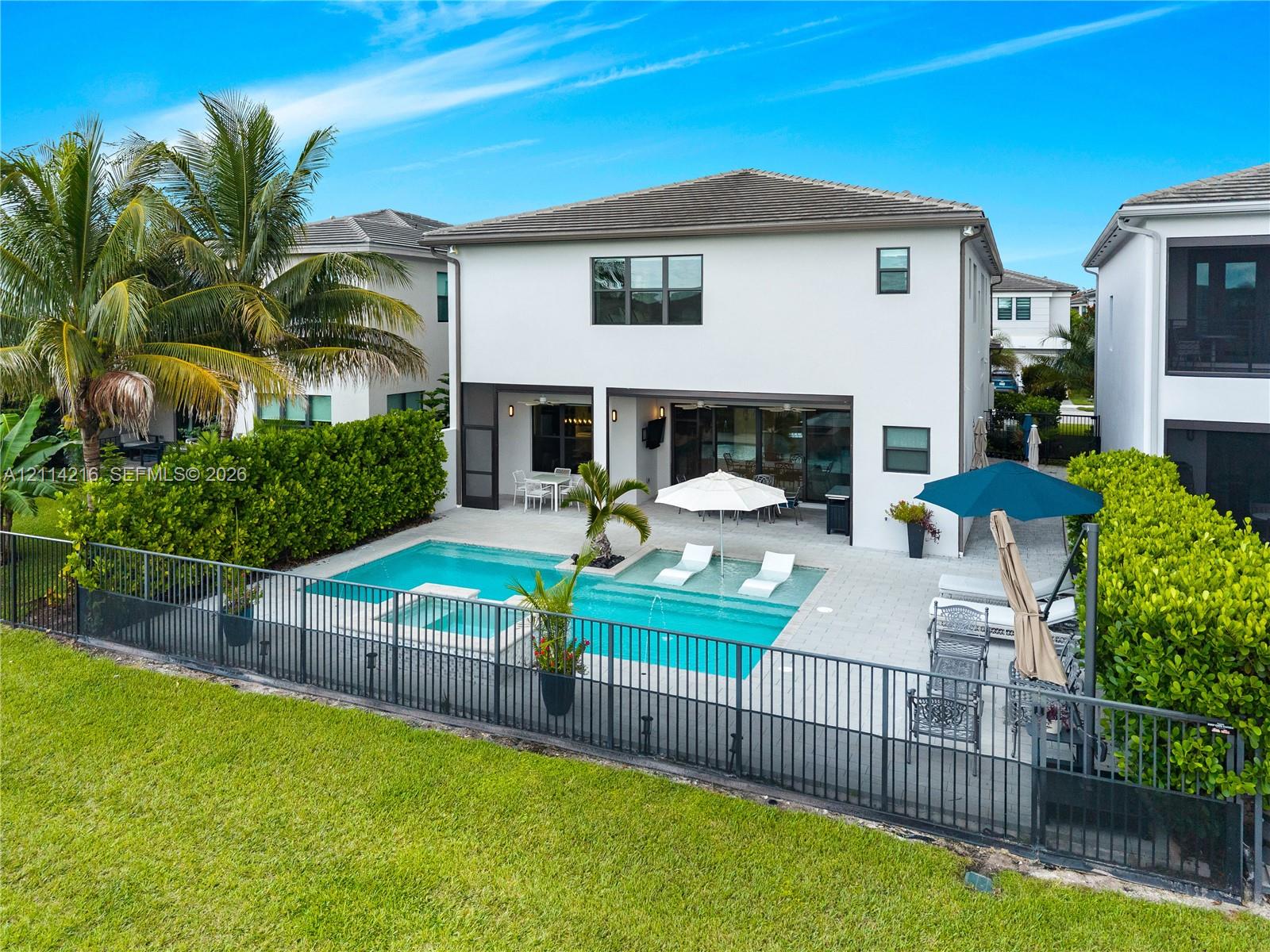 Rear exterior featuring a swimming pool with a sun shelf, integrated spa, paved pool deck, and black aluminum perimeter fencing Rear exterior featuring a swimming pool with a sun shelf, integrated spa, paved pool deck, and black aluminum perimeter fencing