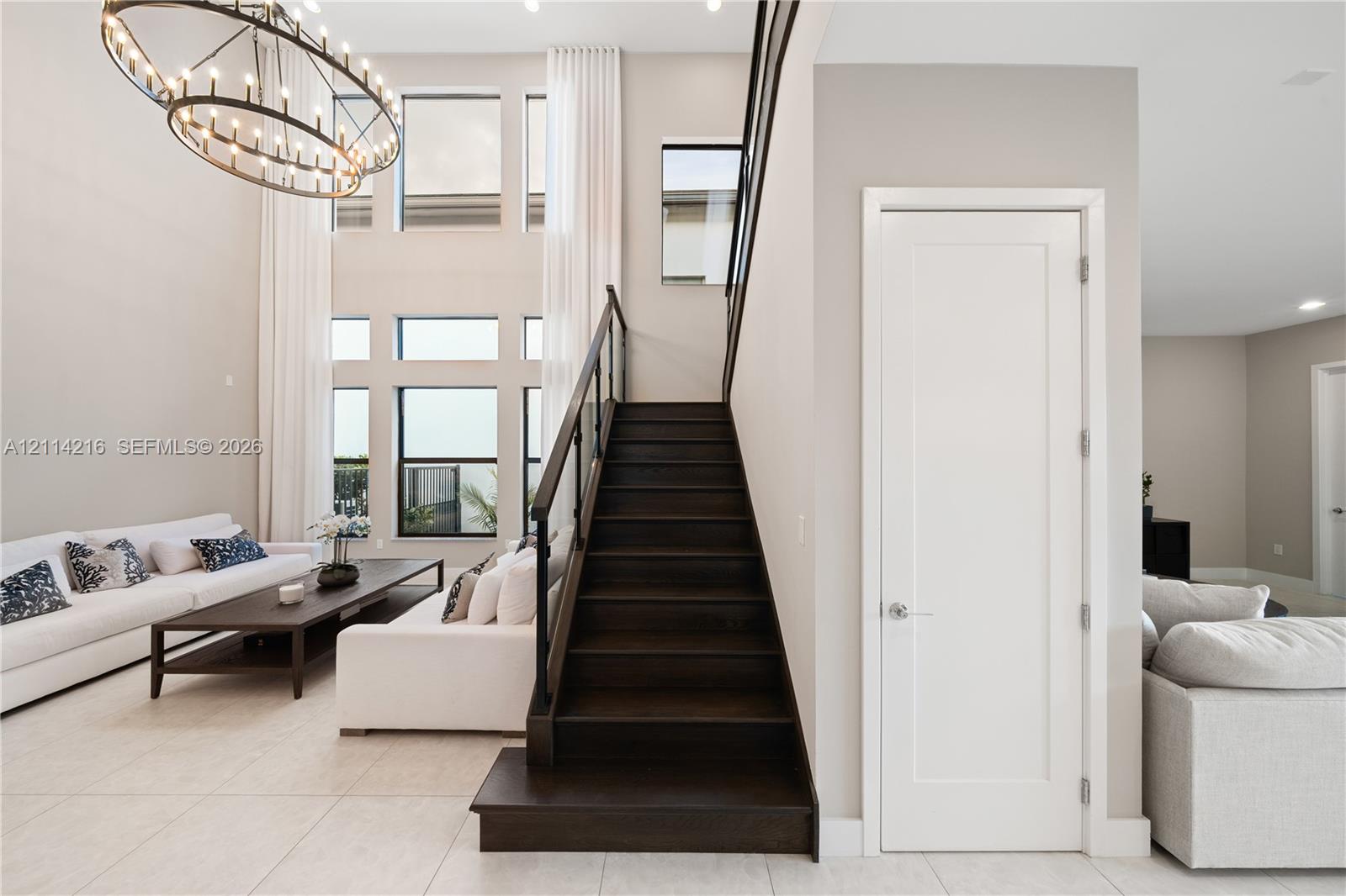 Double-height ceiling, large multi-pane windows, statement ring chandelier, staircase with wood-finish treads and a dark metal railing, and light-toned tile flooring