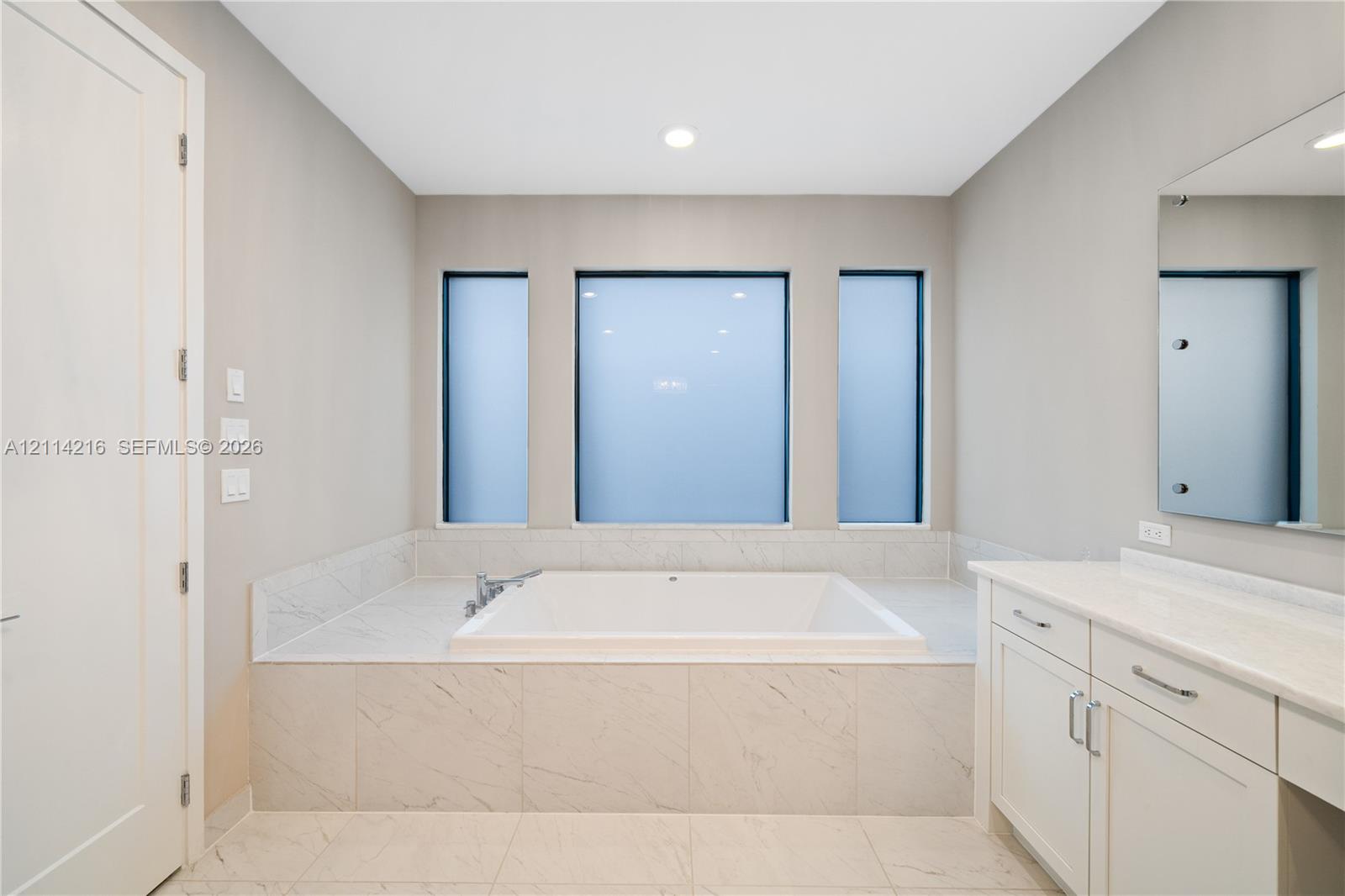 Built-in soaking tub with a tiled surround, triple-window configuration, white cabinetry with stone-finish countertops, and wall-mounted vanity mirror