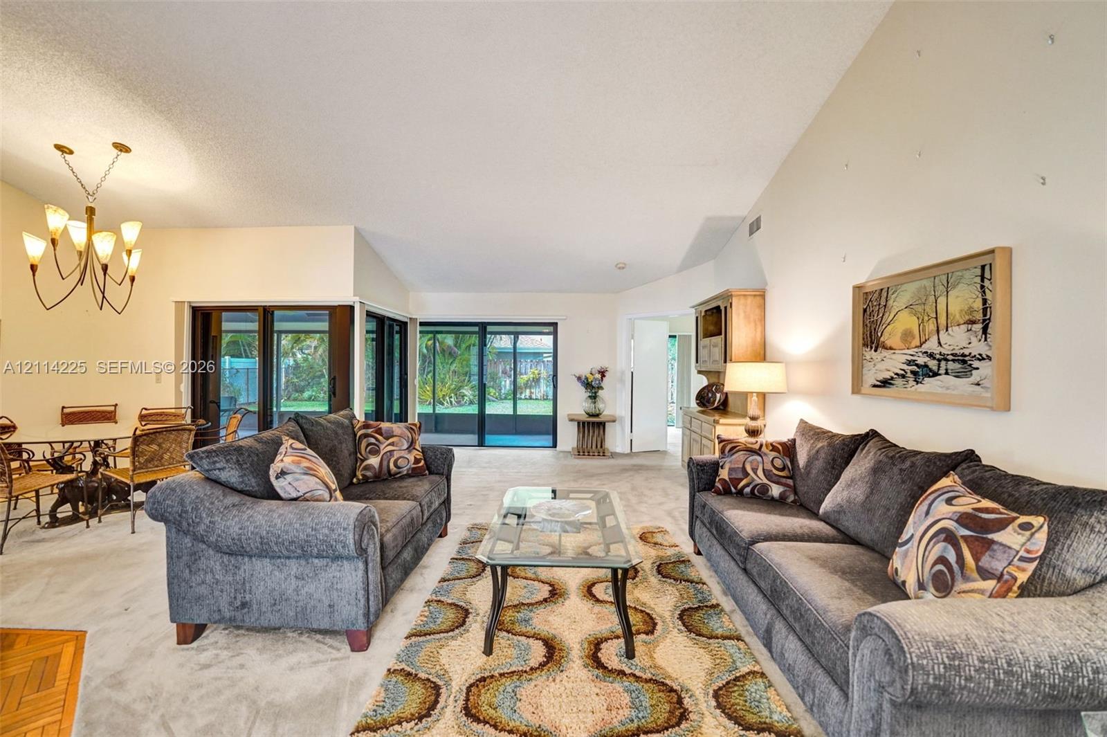 Vaulted ceilings, expansive sliding glass doors, neutral carpeting, and a multi-arm chandelier featuring classic candle-style lighting