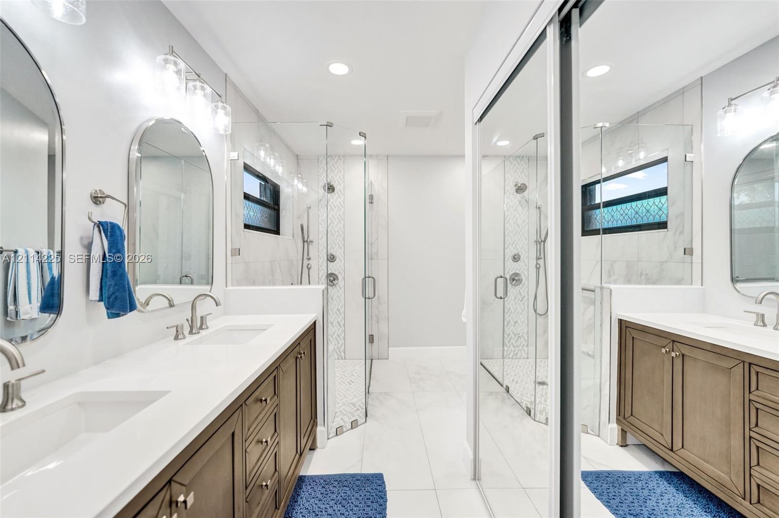 Dual wood-finish vanities with quartz countertops and undermount sinks, arched wall mirrors, and a walk-in glass-enclosed shower featuring stone tile accents and dual showerheads