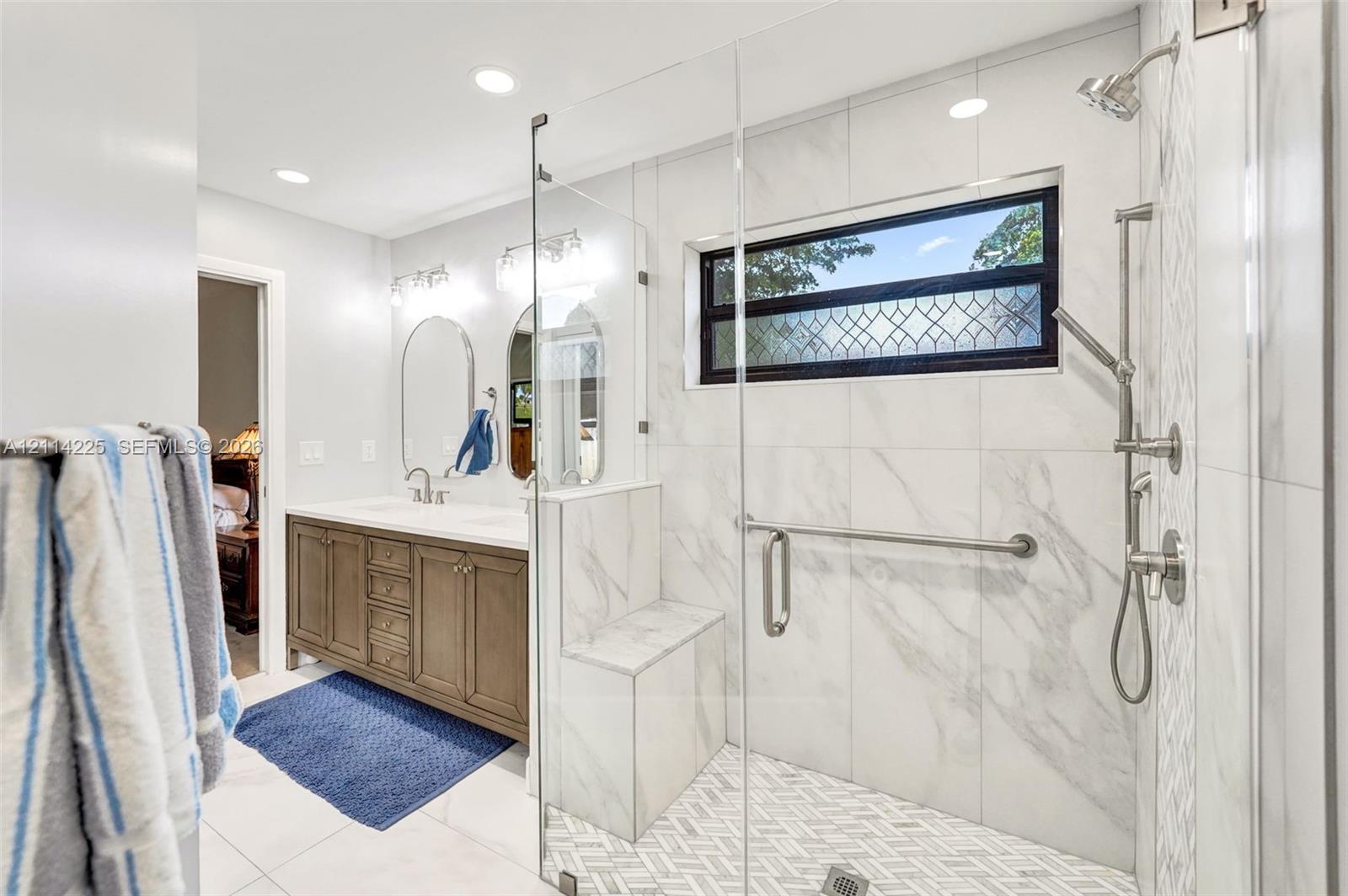 Bathroom featuring a dual-sink vanity, glass-enclosed shower with a tiled bench, herringbone-patterned shower flooring, and wall-mounted plumbing fixtures