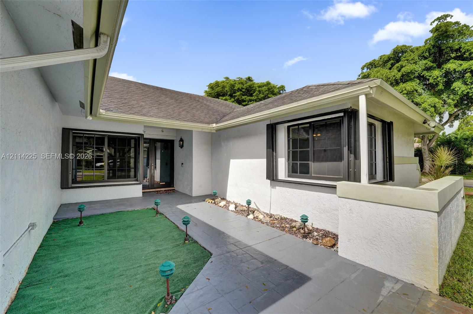 Stucco exterior featuring an asphalt shingle roof, dark-framed grid windows, a covered entryway, and a paved walkway with integrated low-voltage lighting