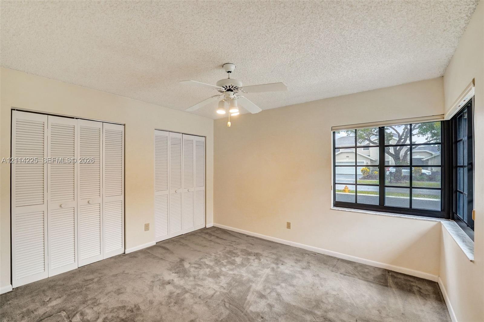 Room featuring neutral carpeting, dual white louvered closet doors, a white ceiling fan with integrated lighting, and a multi-pane corner window