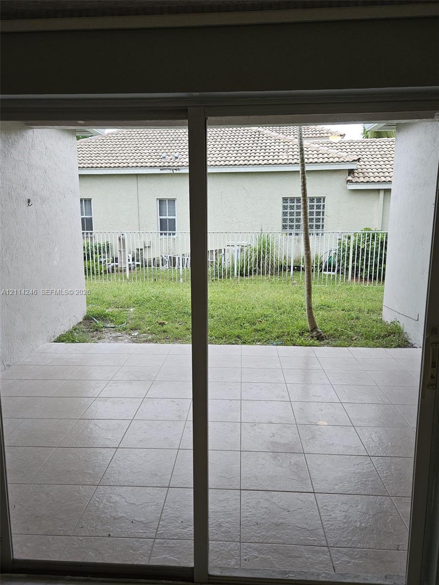Covered patio with neutral-toned square tile flooring, enclosed exterior space with a white metal fence, and grass lawn Covered patio with neutral-toned square tile flooring, enclosed exterior space with a white metal fence, and grass lawn