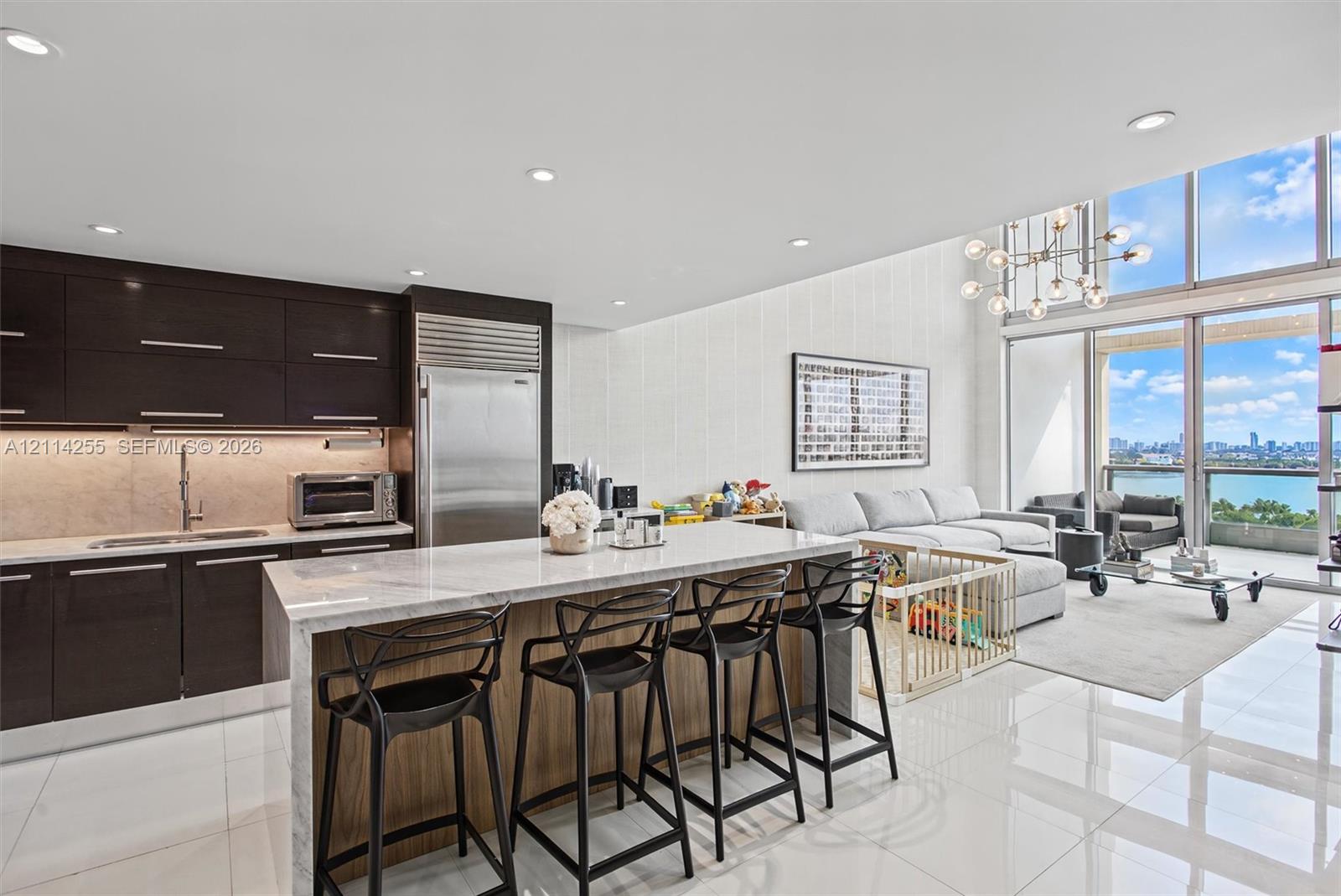 Kitchen featuring dark custom cabinetry, stone-topped waterfall island, professional-grade stainless steel refrigerator, recessed lighting, and floor-to-ceiling windows
