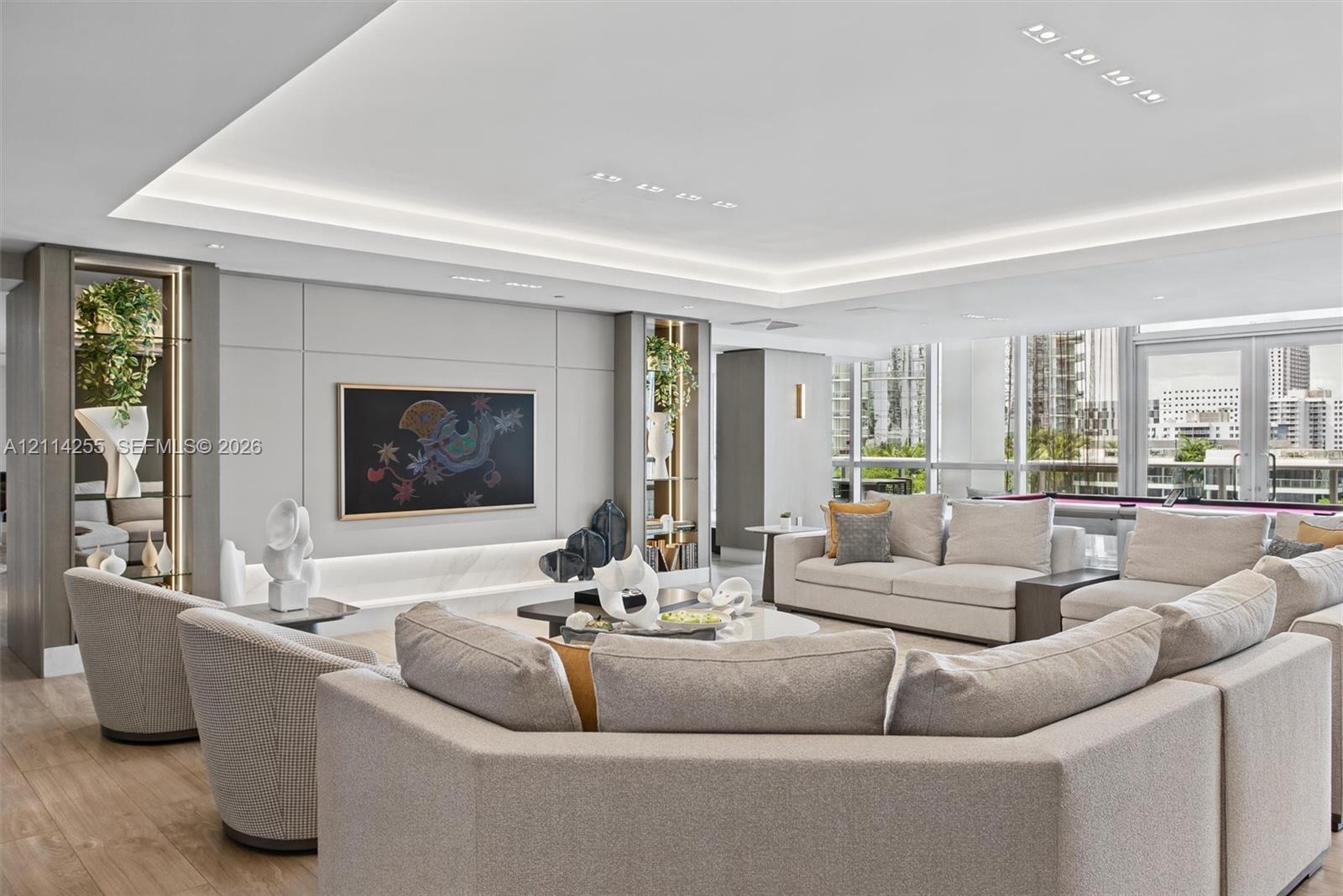 Open-concept living space featuring wood-finish flooring, recessed tray ceiling with integrated cove lighting, floor-to-ceiling glass windows, and built-in shelving units with recessed lighting