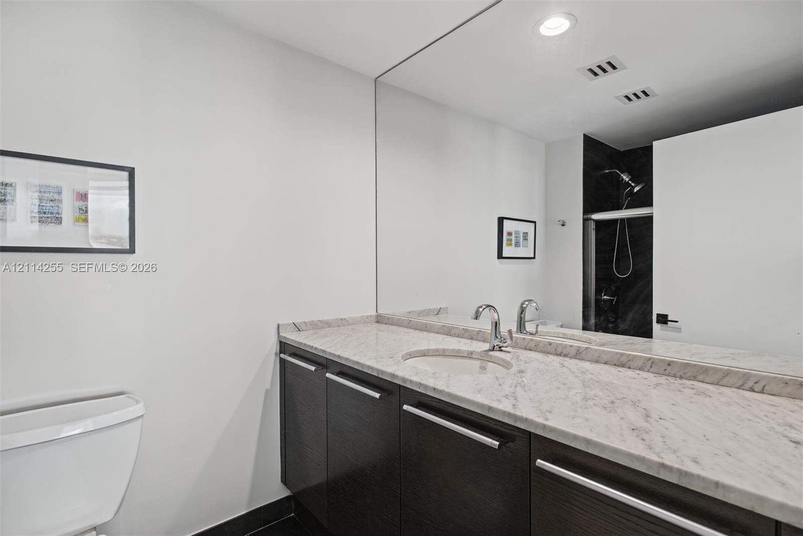 Bathroom featuring dark wood-finish cabinetry, stone countertop with an integrated oval sink, chrome faucet, expansive wall-to-wall mirror, and recessed lighting