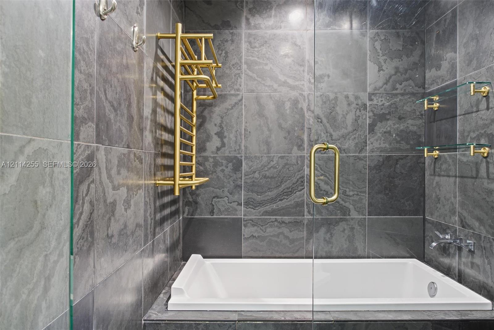 Large-format charcoal stone tile surround with a white deep-soaking tub, brass-finish heated towel rack, wall-mounted faucet, and glass shelving