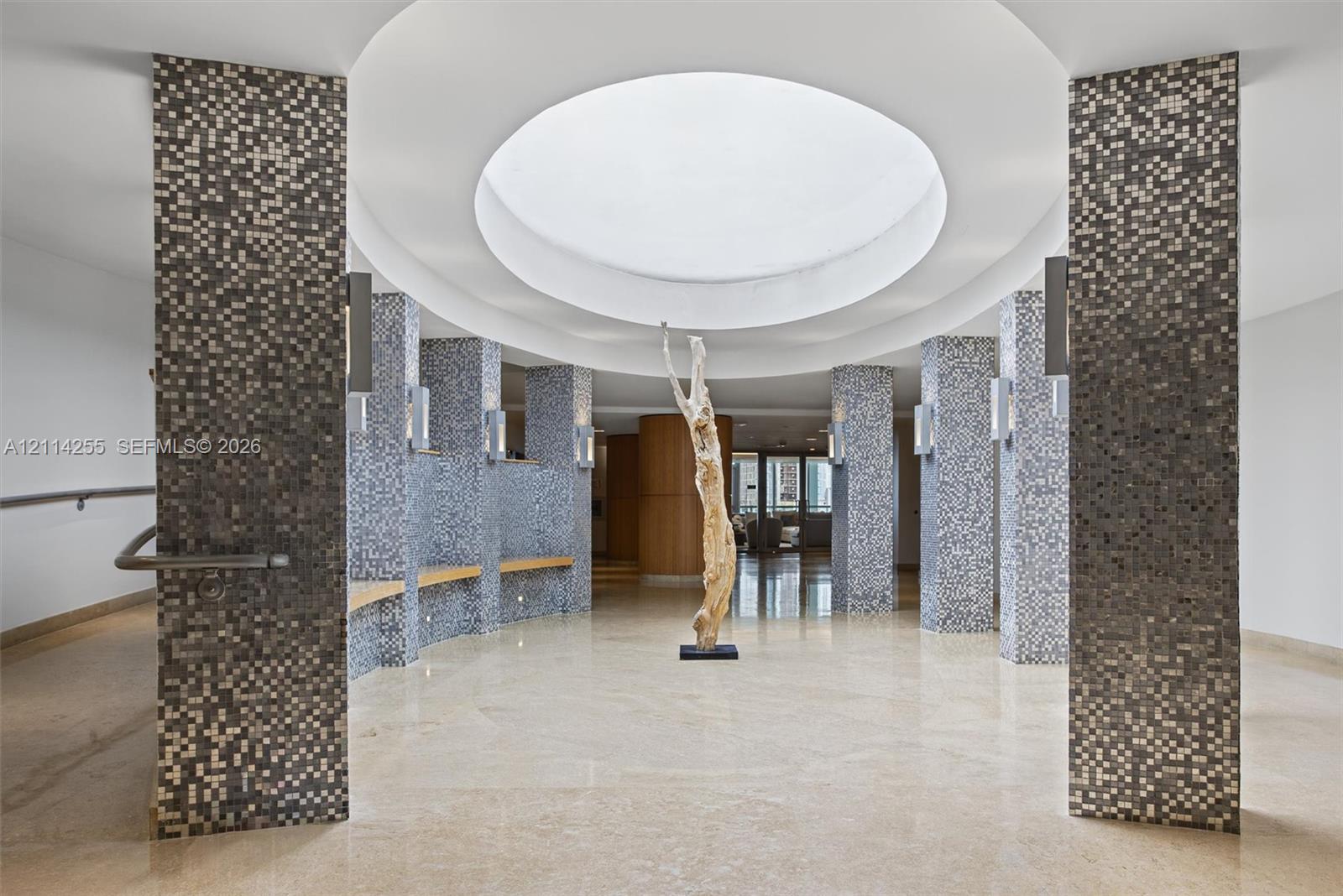 Circular rotunda featuring a recessed dome ceiling, mosaic-tiled structural columns, built-in wooden benches, and polished stone flooring