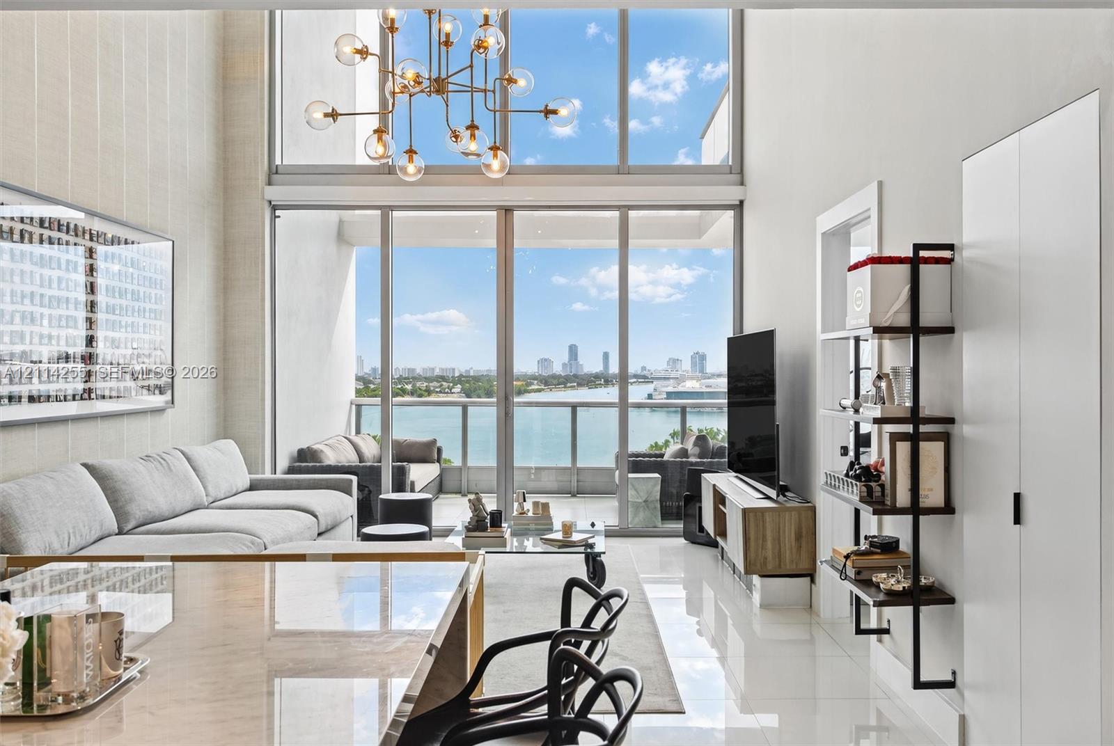 Double-height floor-to-ceiling windows, modern metal chandelier, polished tile flooring, and integrated balcony access with waterfront scenery Double-height floor-to-ceiling windows, modern metal chandelier, polished tile flooring, and integrated balcony access with waterfront scenery