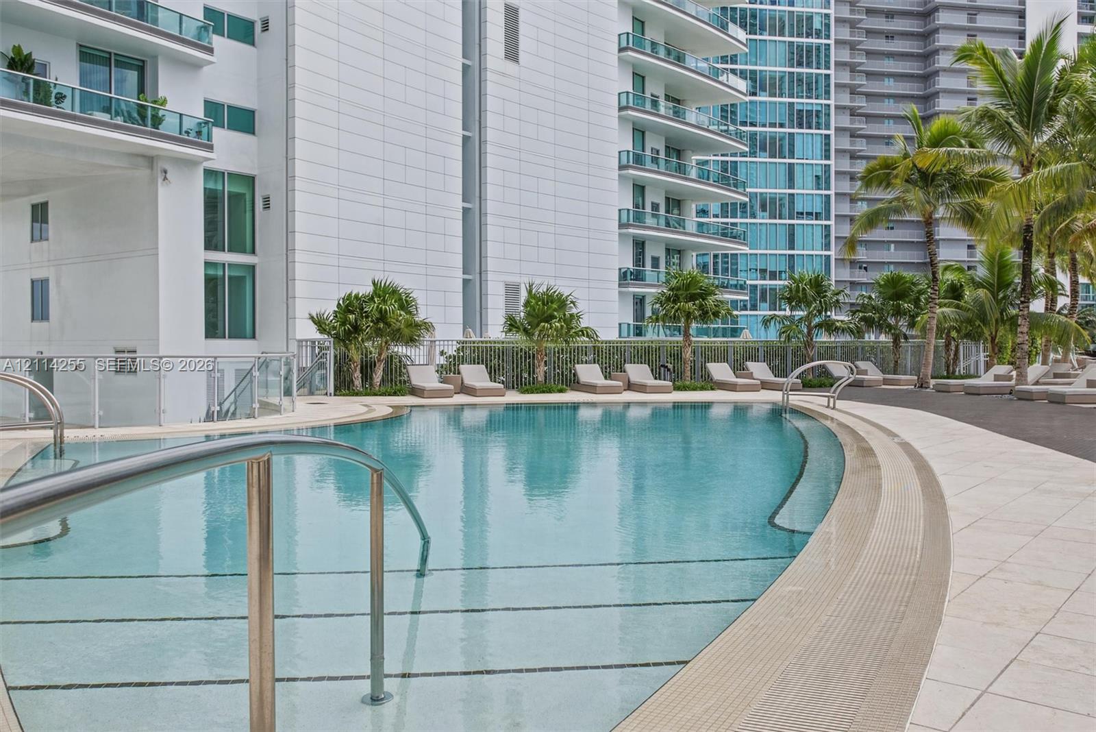 Curved swimming pool featuring a zero-entry design, metal handrails, a tiled deck, and surrounding palm trees