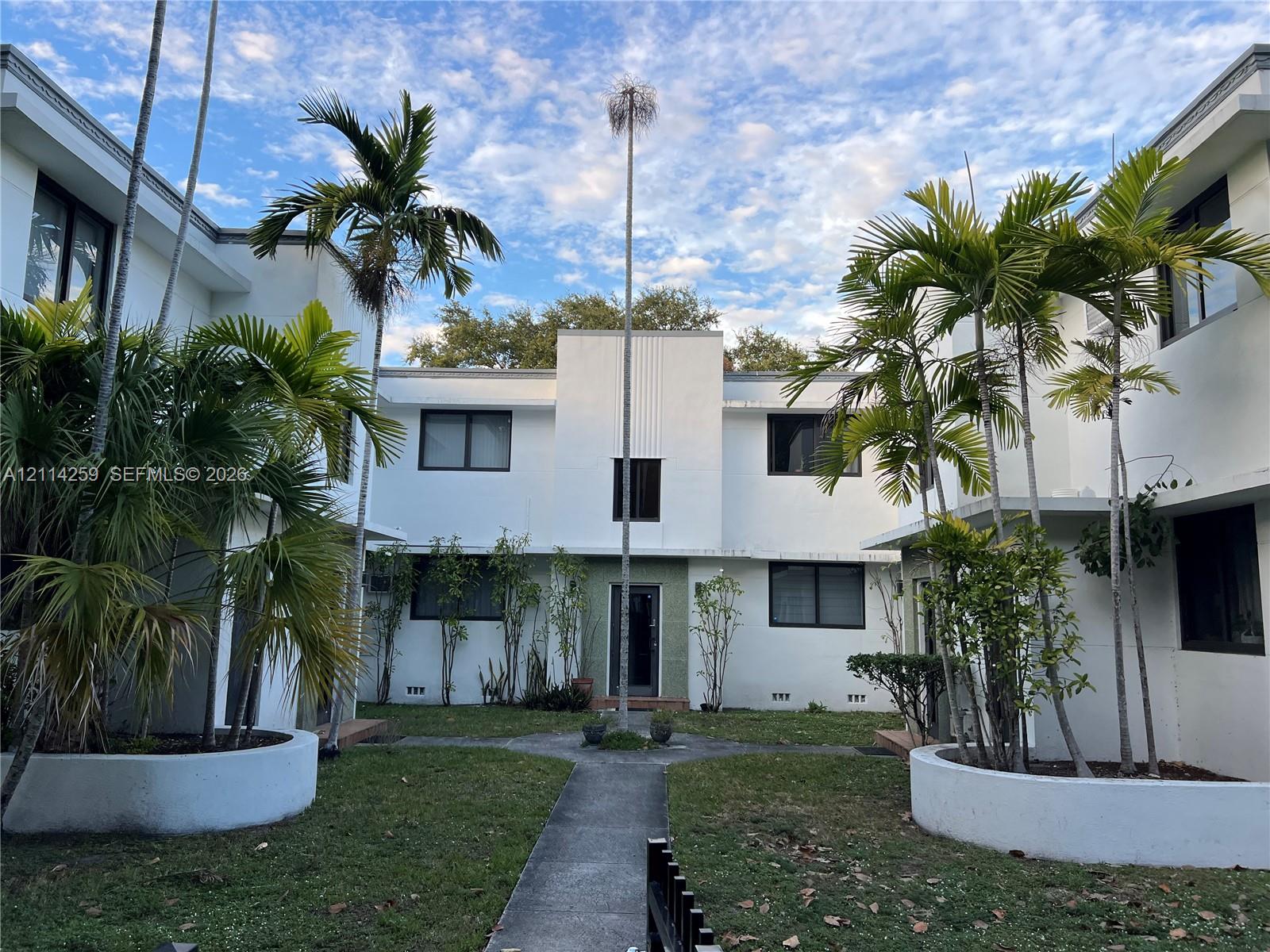 Multi-story stucco exterior featuring a central courtyard with palm trees, concrete pathways, and raised circular planting beds