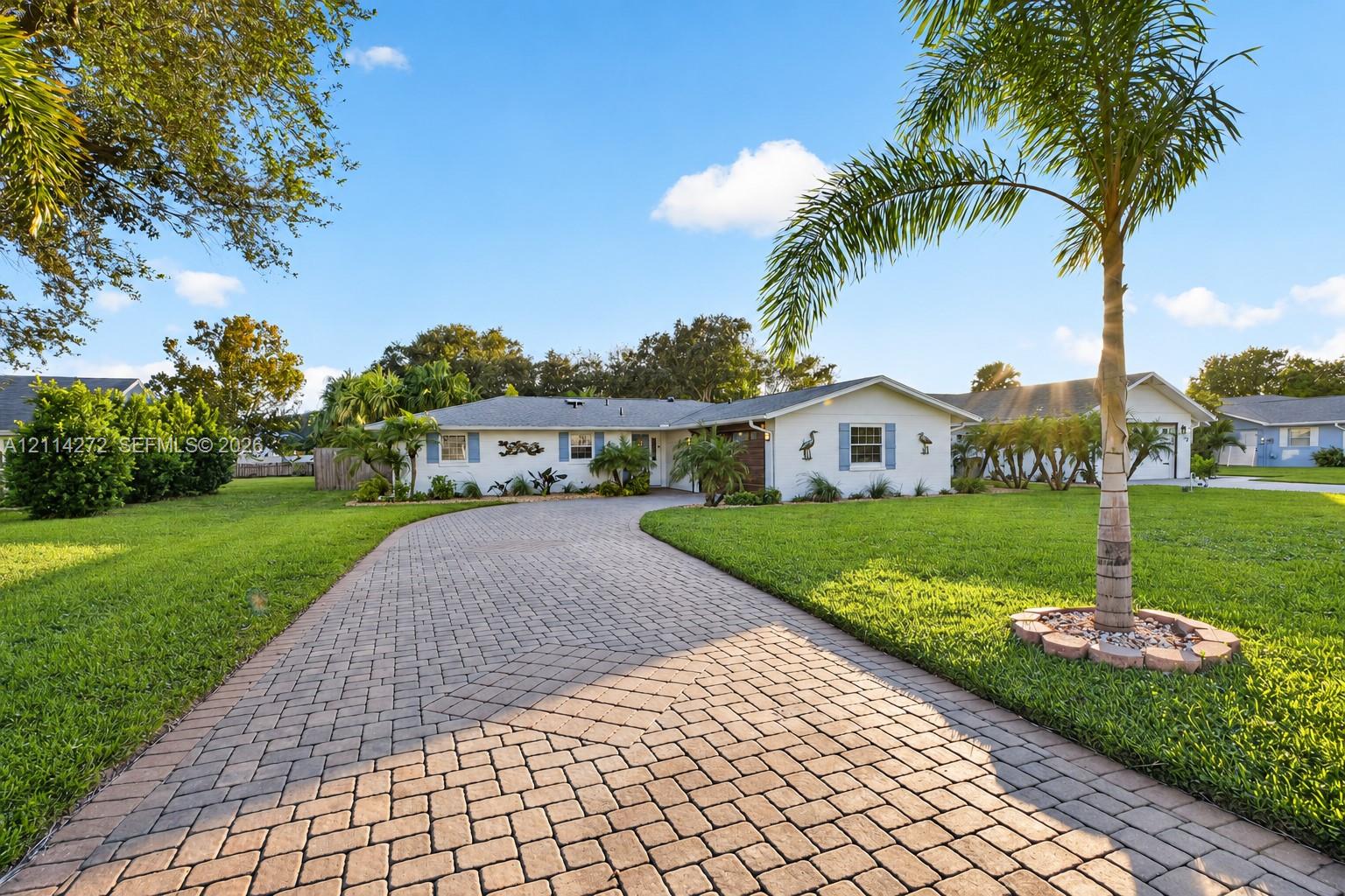 Single-story residence featuring a paved stone driveway, manicured lawn, and mature palm landscaping