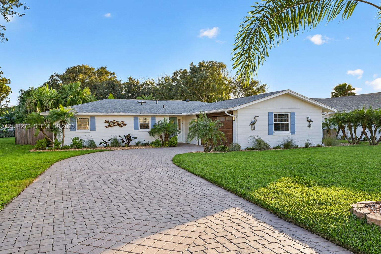 Single-story residence featuring a painted masonry exterior, a wide paver driveway, blue window shutters, and mature landscaping