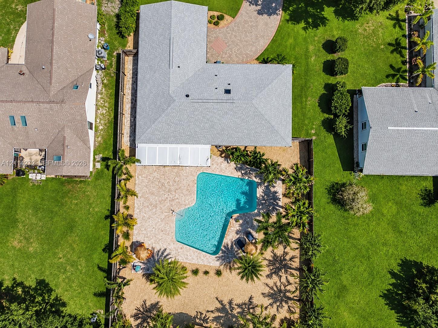 Aerial perspective of a property featuring an in-ground swimming pool, stone-paver pool deck, covered patio, manicured lawn, and paved circular driveway