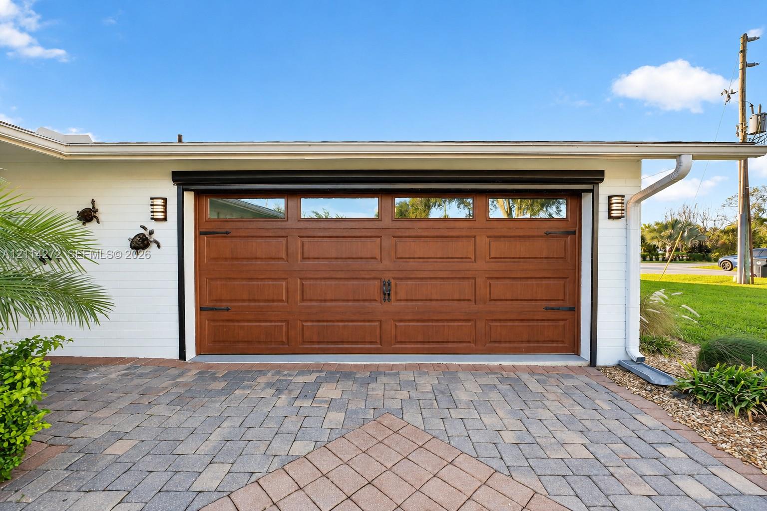 White painted brick exterior featuring a wood-finish sectional garage door with inset windows, decorative black hardware, paver driveway, and mounted exterior wall sconces